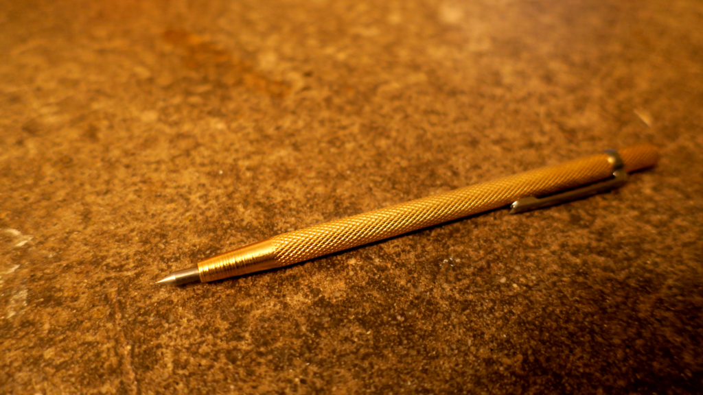 Against a background of concrete, a gold scribe in the shape of a pen with diamond-textured grip. The tip is silvery and sharp. There is a pocket clip at the non-business end.