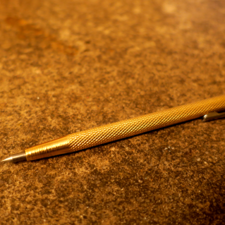 Against a background of concrete, a gold scribe in the shape of a pen with diamond-textured grip. The tip is silvery and sharp. There is a pocket clip at the non-business end.