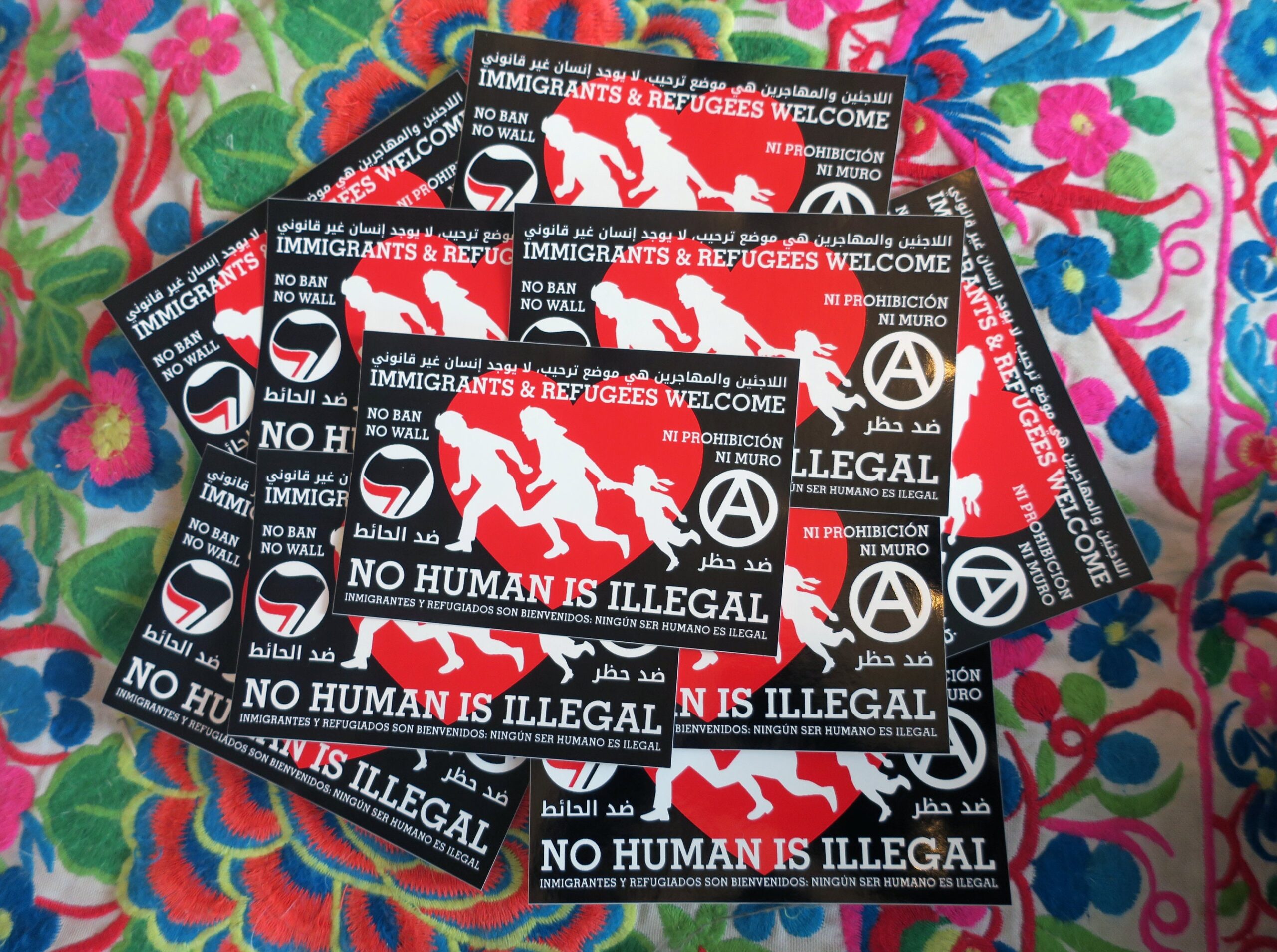 A colorful embroidery, upon which is a pile of 10 stickers: Red heart on a black background. Atop the red heart a silhouetted family of three runs to the right. To the right of the family is an antifa insignia, to the left an anarchist circle A. The words "Immigrants & Refugees Welcome, No Human Is Illegal, No Ban No Wall," "ﺍﻟﻼﺟﺌﻴﻦ ﻭﺍﻟﻤﻬﺎﺟﺮﻳﻦ ﻫﻲ ﻣﻮﺿﻊ ﺗﺮﺣﻴﺐ، ﻻ ﻳﻮﺟﺪ ﺇﻧﺴﺎﻥ ﻏﻴﺮ ﻗﺎﻧﻮﻧﻲ, ﺿﺪ ﺍﻟﺤﺎﺋﻂ ﺿﺪ ﺣﻈﺮ" and "Inmigrantes y refugiados son bienvenidos, Ningún ser humano es ilegal, Ni prohibición ni muro" appear around the heart.