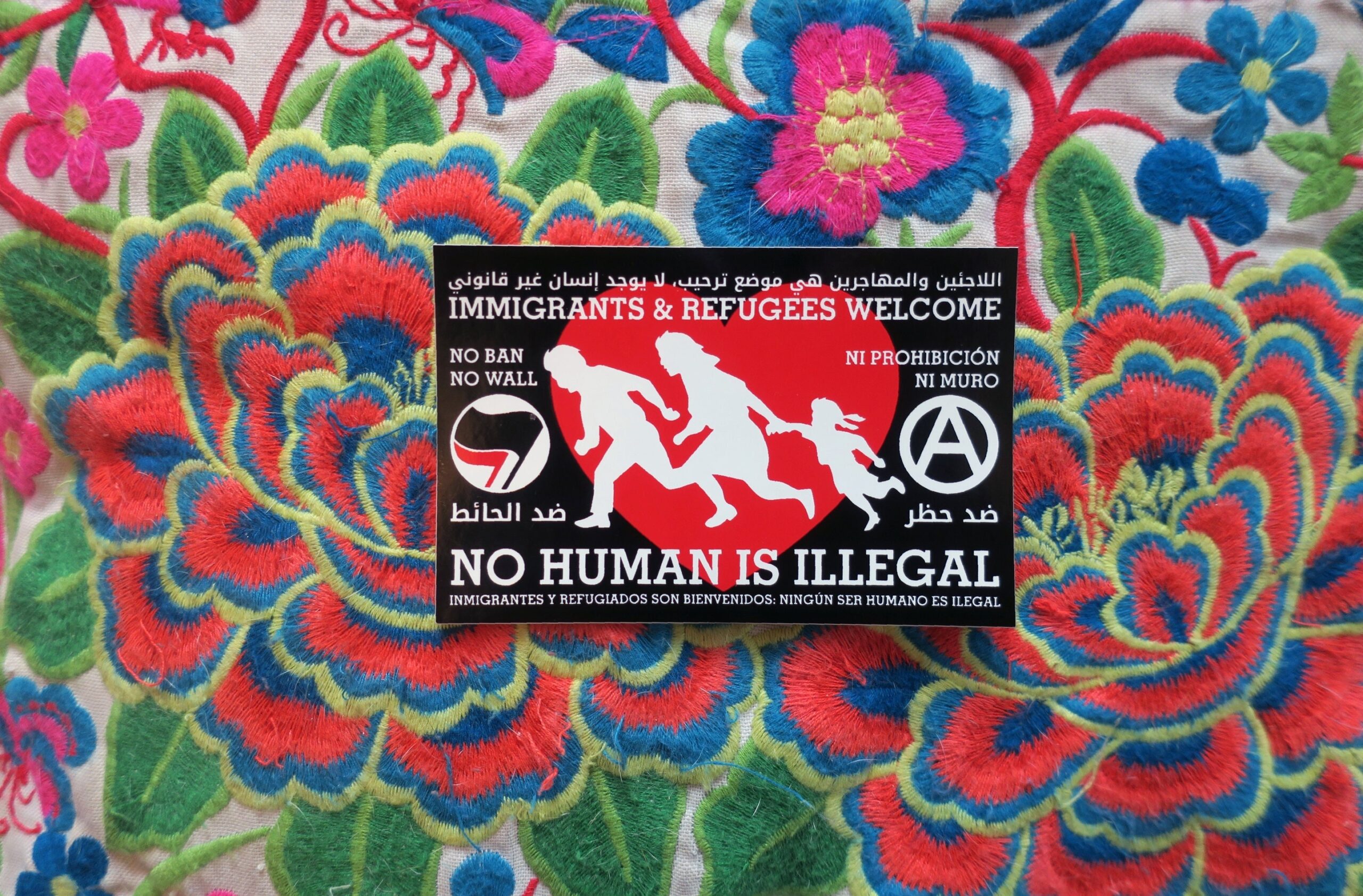 A colorful embroidery, upon which is a sticker: Red heart on a black background. Atop the red heart a silhouetted family of three runs to the right. To the right of the family is an antifa insignia, to the left an anarchist circle A. The words "Immigrants & Refugees Welcome, No Human Is Illegal, No Ban No Wall," "ﺍﻟﻼﺟﺌﻴﻦ ﻭﺍﻟﻤﻬﺎﺟﺮﻳﻦ ﻫﻲ ﻣﻮﺿﻊ ﺗﺮﺣﻴﺐ، ﻻ ﻳﻮﺟﺪ ﺇﻧﺴﺎﻥ ﻏﻴﺮ ﻗﺎﻧﻮﻧﻲ, ﺿﺪ ﺍﻟﺤﺎﺋﻂ ﺿﺪ ﺣﻈﺮ" and "Inmigrantes y refugiados son bienvenidos, Ningún ser humano es ilegal, Ni prohibición ni muro" appear around the heart.