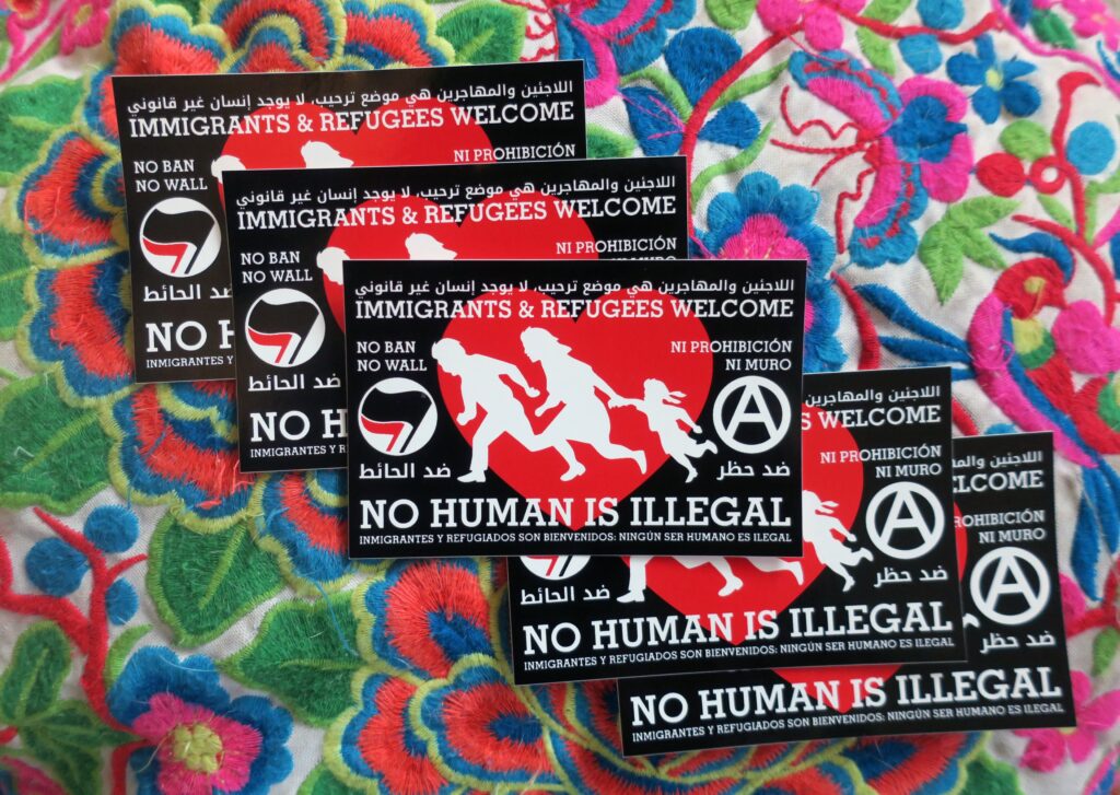 A colorful embroidery, upon which is a cascade of 5 stickers: Red heart on a black background. Atop the red heart a silhouetted family of three runs to the right. To the right of the family is an antifa insignia, to the left an anarchist circle A. The words "Immigrants & Refugees Welcome, No Human Is Illegal, No Ban No Wall," "ﺍﻟﻼﺟﺌﻴﻦ ﻭﺍﻟﻤﻬﺎﺟﺮﻳﻦ ﻫﻲ ﻣﻮﺿﻊ ﺗﺮﺣﻴﺐ، ﻻ ﻳﻮﺟﺪ ﺇﻧﺴﺎﻥ ﻏﻴﺮ ﻗﺎﻧﻮﻧﻲ, ﺿﺪ ﺍﻟﺤﺎﺋﻂ ﺿﺪ ﺣﻈﺮ" and "Inmigrantes y refugiados son bienvenidos, Ningún ser humano es ilegal, Ni prohibición ni muro" appear around the heart.