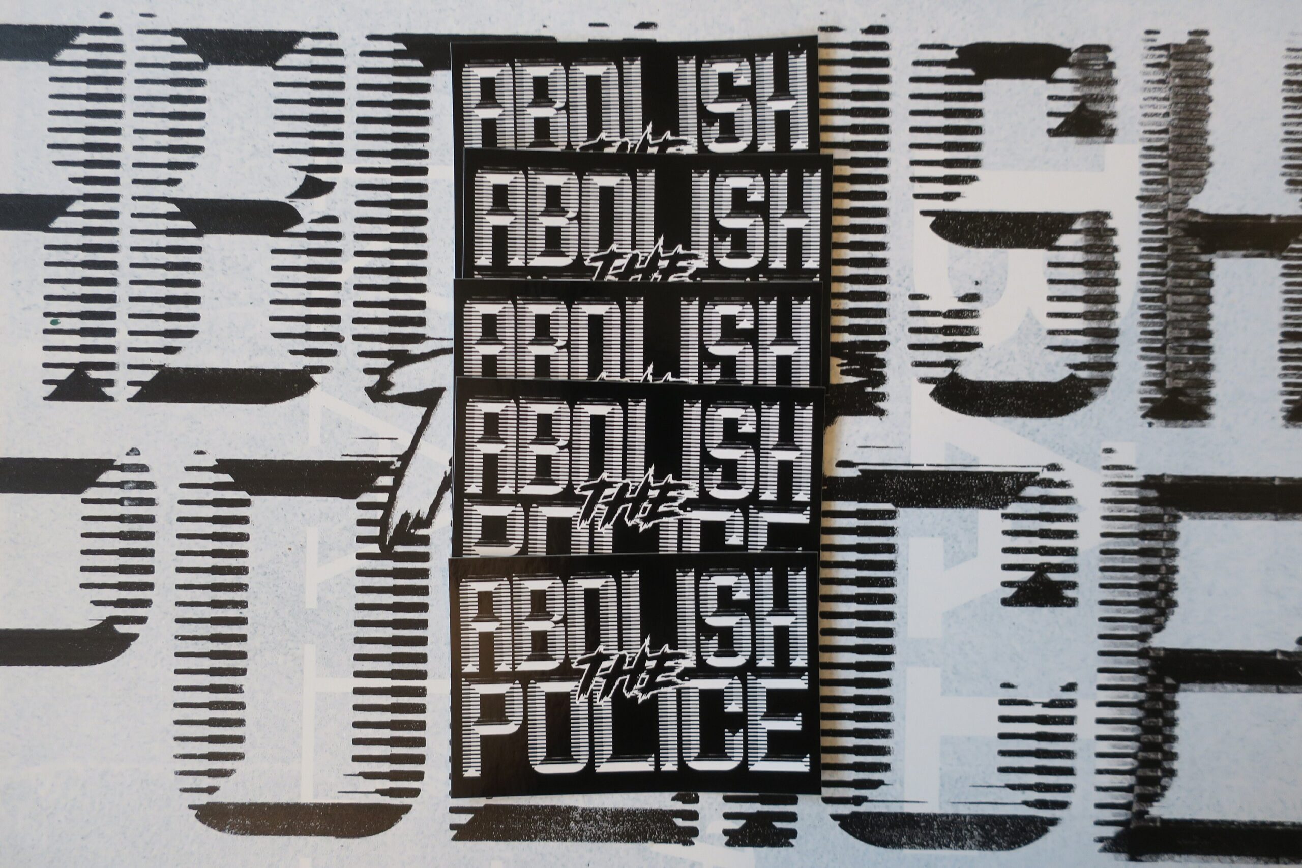 A background of a white sign with black retrofuturistic typeface reading "Abolish the Police." partially obscured by 5 black stickers with white retrofuturistic typeface reading "Abolish the Police."