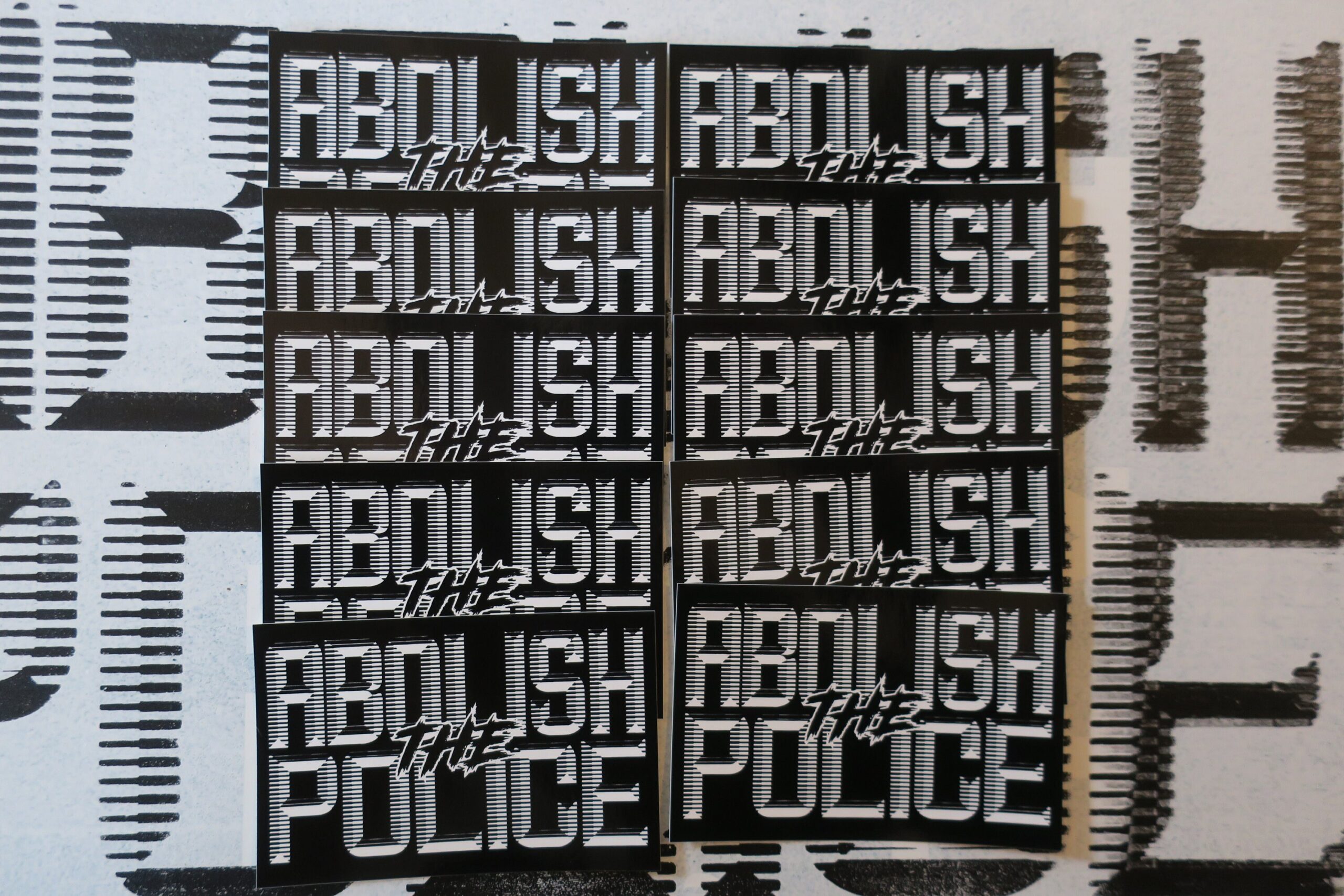 A background of a white sign with black retrofuturistic typeface reading "Abolish the Police." partially obscured by 10 black stickers in two columns with white retrofuturistic typeface reading "Abolish the Police."