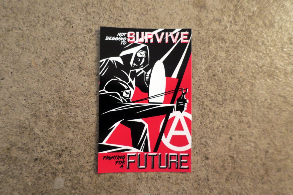 On a background of concrete, a sticker printed with a black and white cartoon depiction of two front line rioters in hoods, respirators, goggles and black clothing. One of them crouches wielding a slingshot pulled ready to fire. The other braces behind a shield. In front of and above both rioters waves a black flag. Behind the rioters, a white circle-a anarchy symbol. Superimposed on top of these rioters, in retrofuturistic typeface, are the words, "Not begging to survive, fighting for a future" in black, red and white.