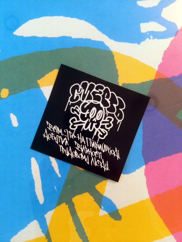 A colorful silkscreen backdrop. Laid atop this is a black square sticker: Bubble letters arc across the top, the Ukrainian word Смерть, or DEATH. Below this word is a graffiti stylized skull and crossbones, looking to the right. There are drips of paint coming down off of the bubble letters and the skull. Below the skull, in graffiti handstyle are the words всім, хто на пиришкоді добутья вільності трудовому люду, or "to those who stand in the way of freedom for working people."