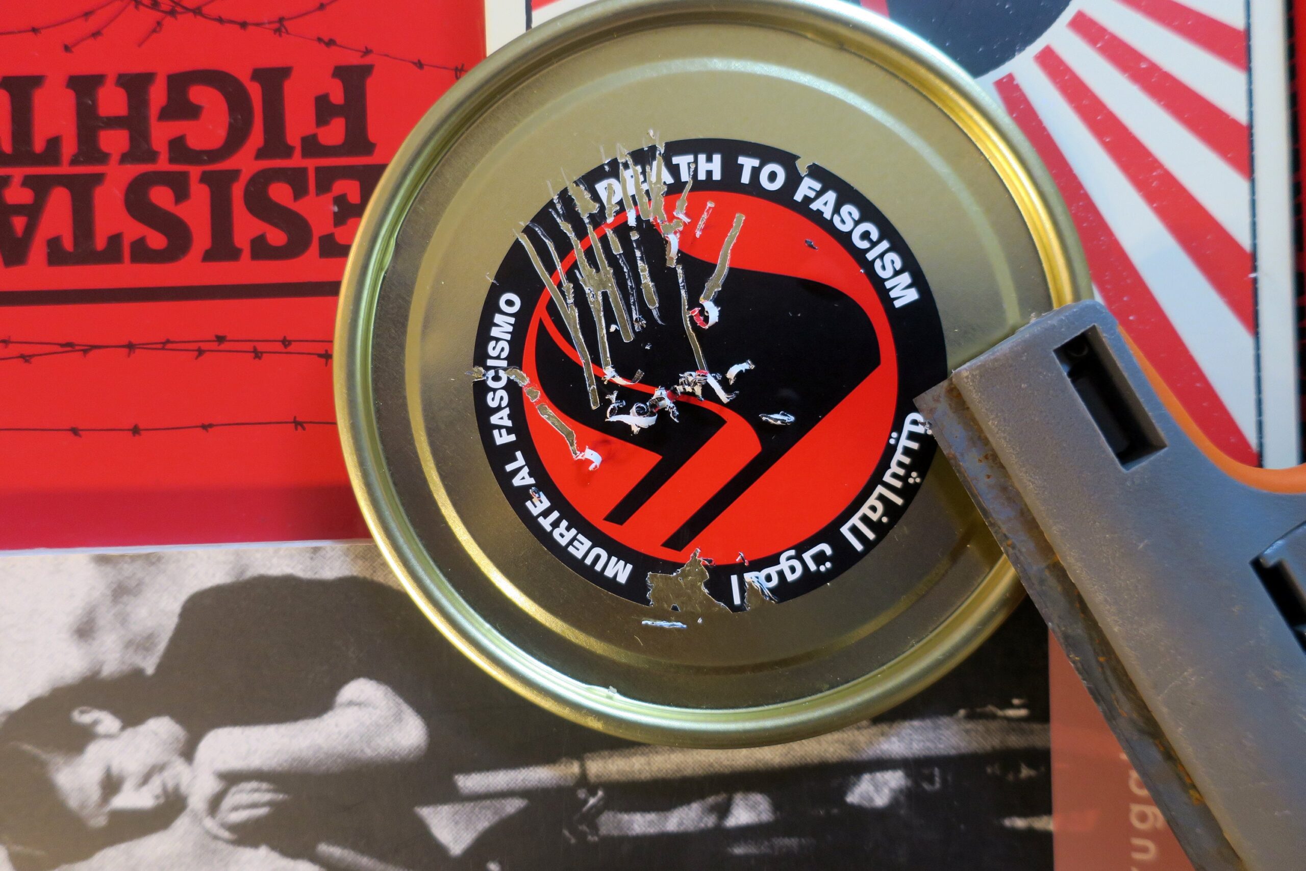 On a backdrop of book covers including Antifa: the Antifascist Handbook, Free Women of Spain, and Resistance Fighter, metal can lid with a sticker affixed to it. The sticker is a thick black circle containing the words: "Death to Fascism, Muerte al Fascismo, الموت للفاشية,." Inside this circle are two black flags arranged over a red field recognizable as the antifascist logo. The sticker has been peeled away in thin strips but is entirely recognizable despite efforts the efforts of a razor-edged paint scraper, which also rests on the can lid.