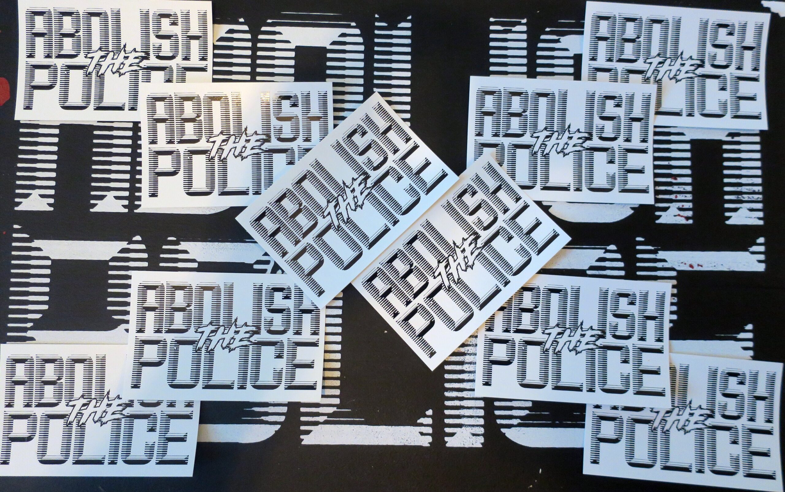 A background of a black sign with white retrofuturistic typeface reading "Abolish the Police." partially obscured by 10 white stickers arranged in an X with black retrofuturistic typeface reading "Abolish the Police."