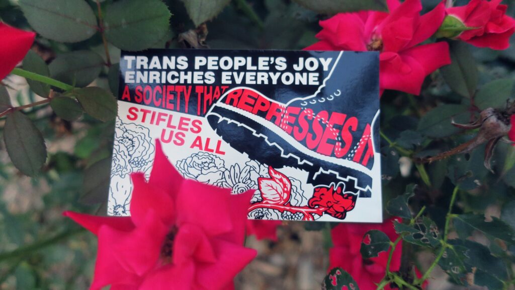 An overripe red rosebush with a sticker embedded between its flowers: A line drawing of a bouquet of flowers with a single red flower being crushed by a large black jackboot. The words, "Trans People's Joy Enriches Everyone, A Society that Represses It Stifles Us All" appear above, through, and just below the boot.