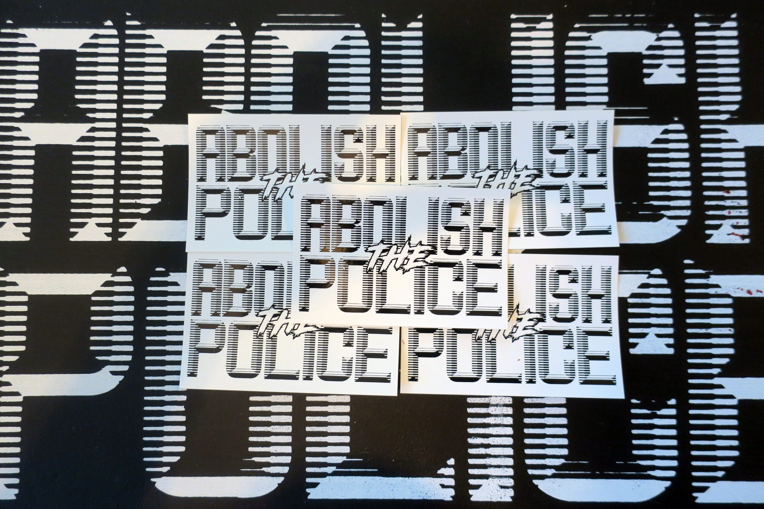 A background of a black sign with white retrofuturistic typeface reading "Abolish the Police." partially obscured by 5 white stickers with black retrofuturistic typeface reading "Abolish the Police."