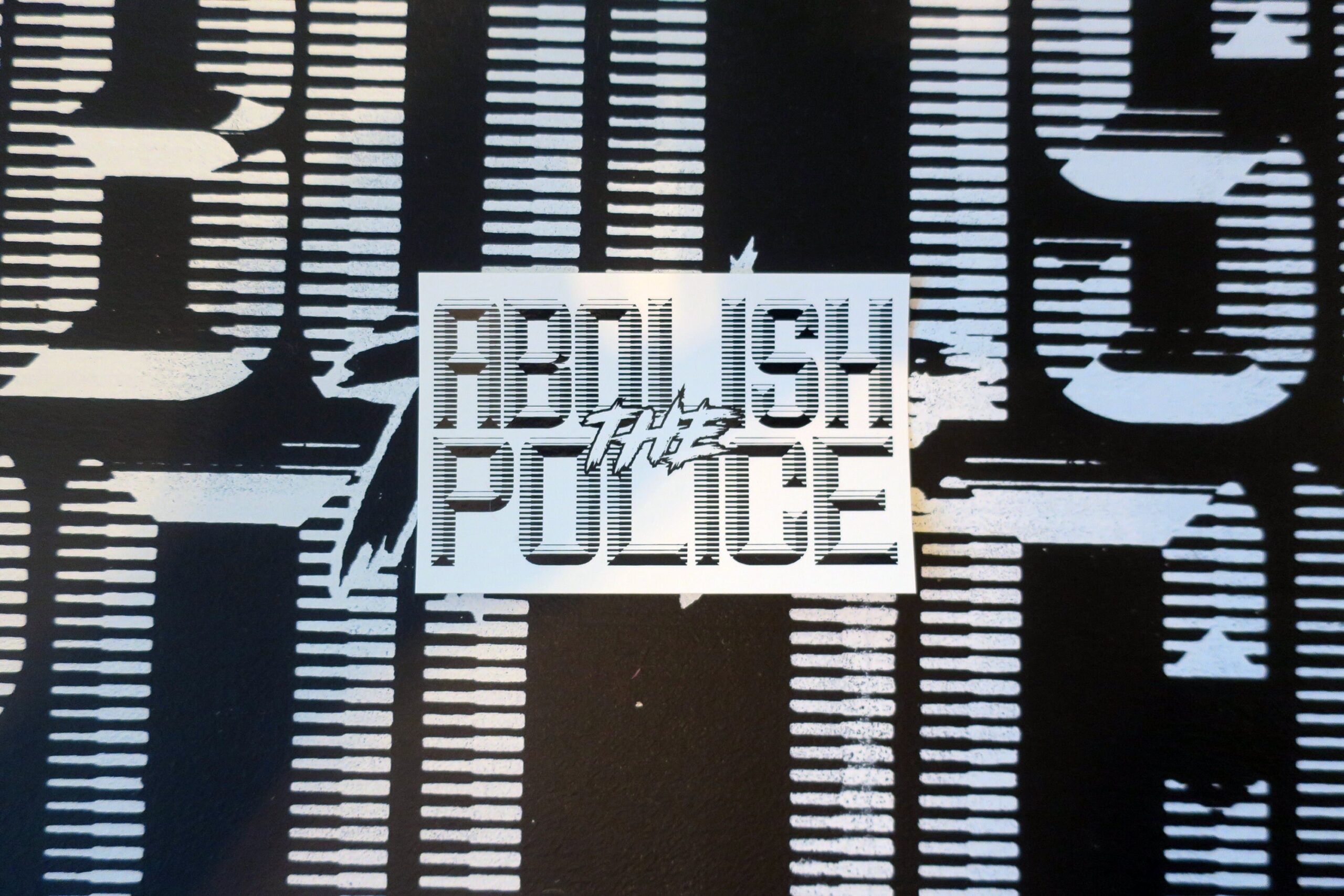 A background of a black sign with white retrofuturistic typeface reading "Abolish the Police." partially obscuredby the cropped picture, upon which is a white sticker with black retrofuturistic typeface reading "Abolish the Police."