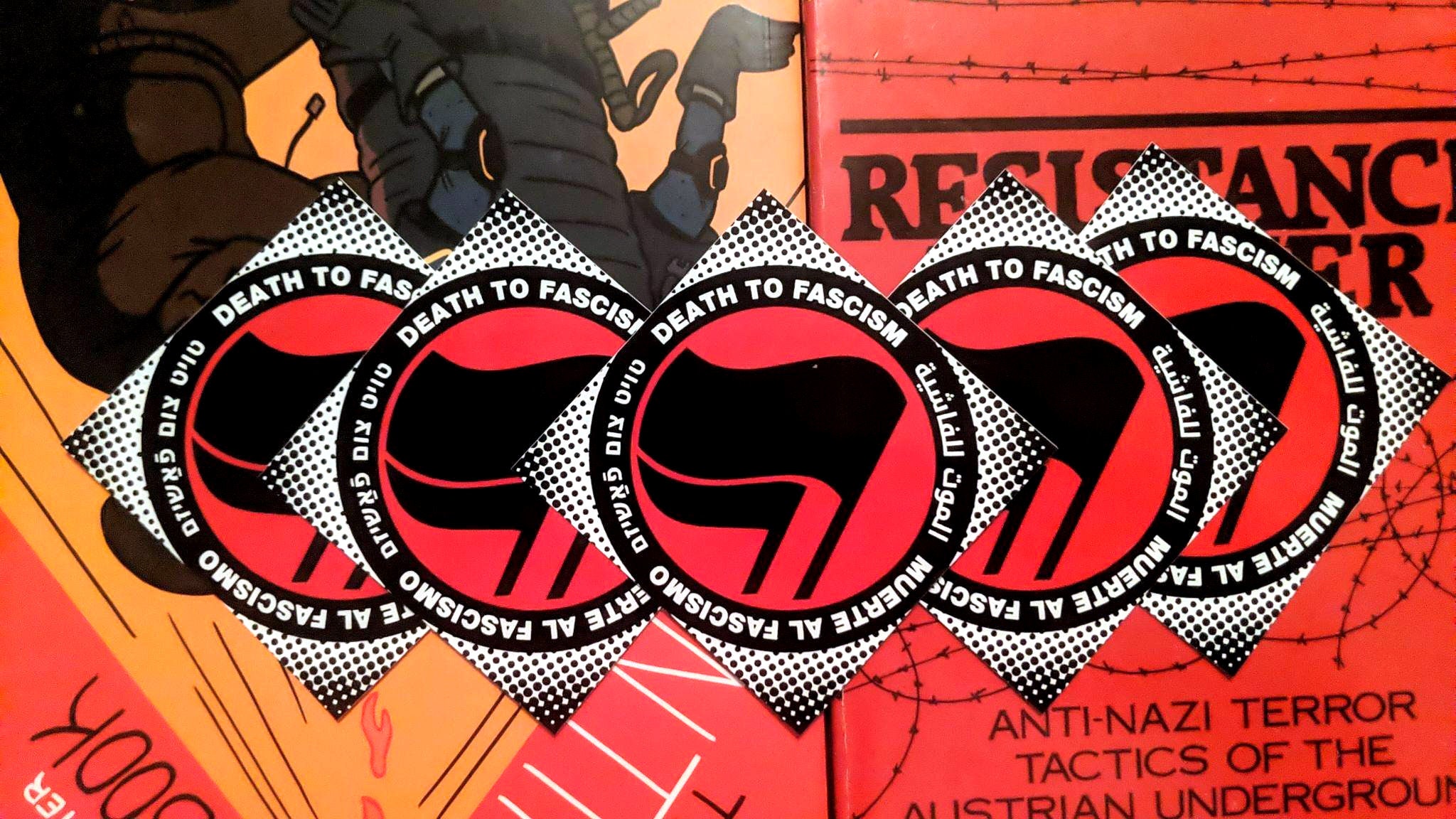 A tidy row of diamond shaped stickers atop a variety of antifascist books. The sticker consists of a halftone halo surrounding a thick black circle with white words: "Death to Fascism, Muerte al Fascismo, الموت للفاشية . טויט צום פֿאַשיזם" Inside this circle are two black antifascist flags over a red field.