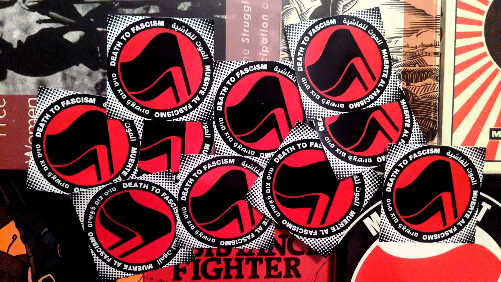 A sloppy pile of diamond shaped stickers atop a variety of antifascist books. The sticker consists of a halftone halo surrounding a thick black circle with white words: "Death to Fascism, Muerte al Fascismo, الموت للفاشية . טויט צום פֿאַשיזם" Inside this circle are two black antifascist flags over a red field.
