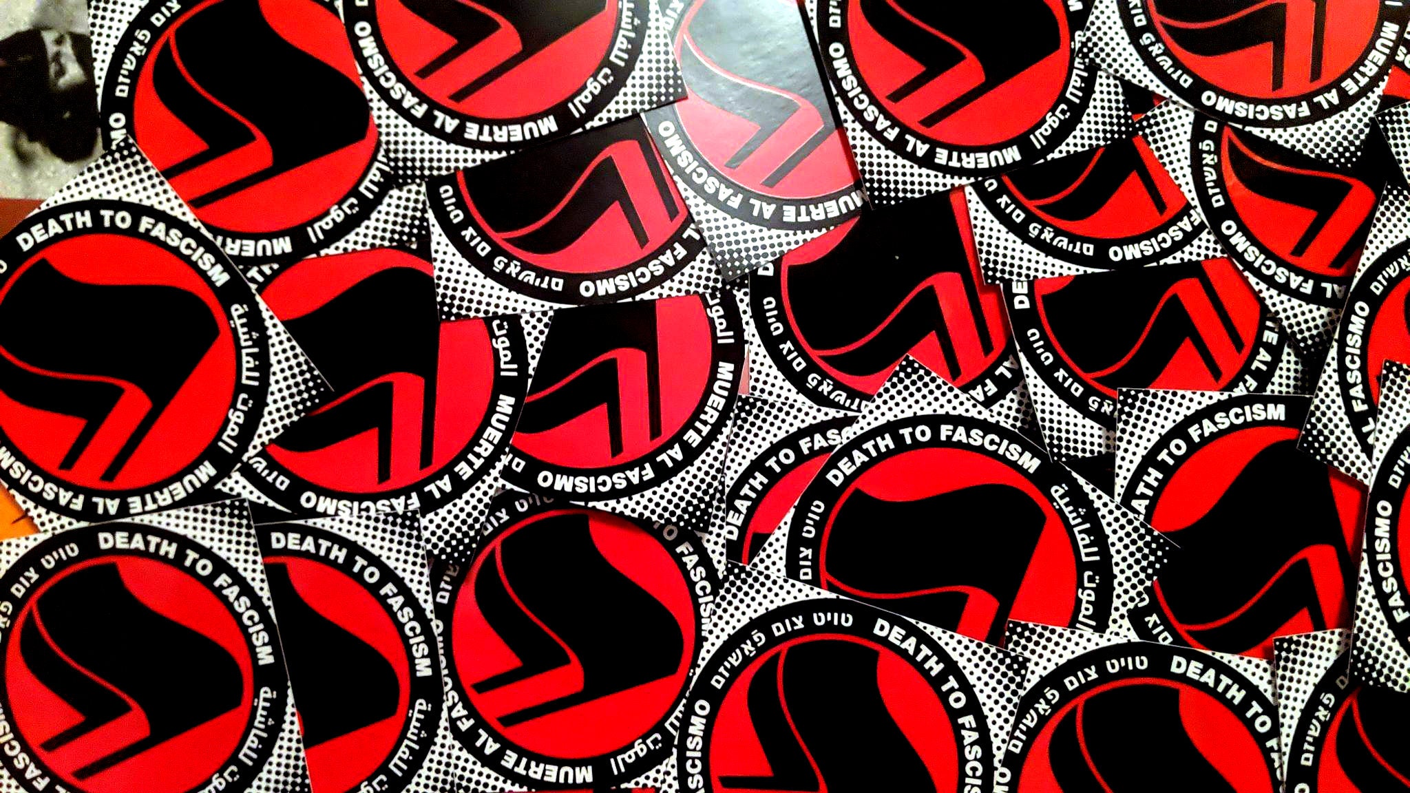 A sloppy pile of diamond shaped stickers (a miliciana of the Free Women of Spain peeks over the top left corner). The sticker consists of a halftone halo surrounding a thick black circle with white words: "Death to Fascism, Muerte al Fascismo, الموت للفاشية . טויט צום פֿאַשיזם" Inside this circle are two black antifascist flags over a red field.