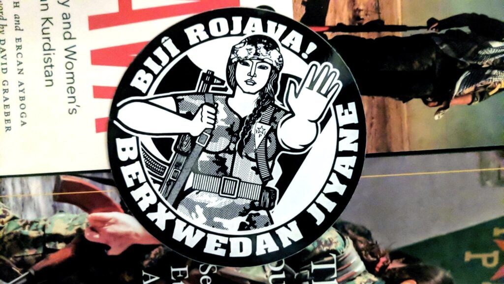On a pile of books, a circular sticker: "Biji Rojava!" arced across the top and "Berxwedan Jiyane" across the bottom. At the center, a stylized image of a Kurdish YPJ militiawoman holding an AK in her right arm and her outstretched hand in a "stop" gesture with her left.