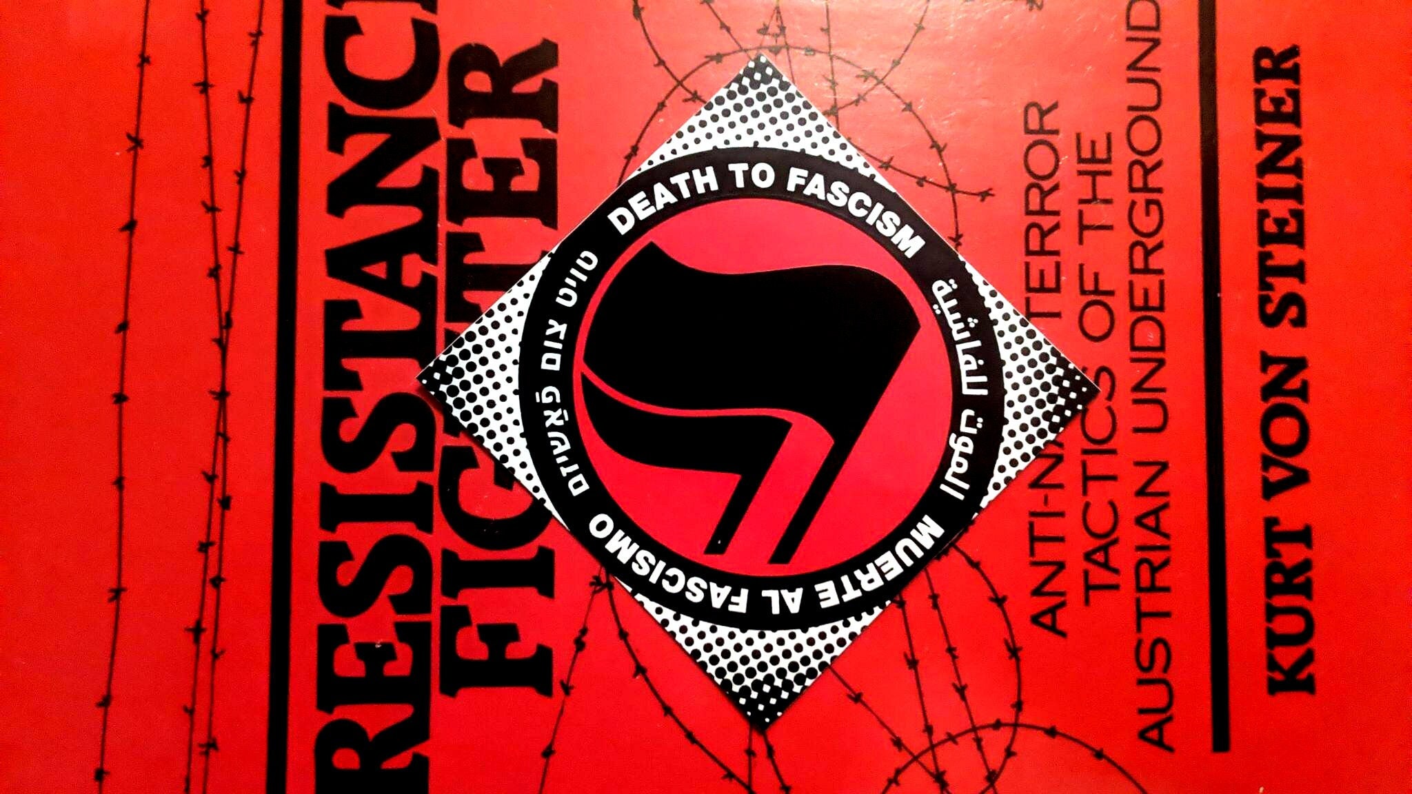 A diamond shaped sticker atop a red book titled Resistance Fighter. The sticker consists of a halftone halo surrounding a thick black circle with white words: "Death to Fascism, Muerte al Fascismo, الموت للفاشية . טויט צום פֿאַשיזם" Inside this circle are two black antifascist flags over a red field.