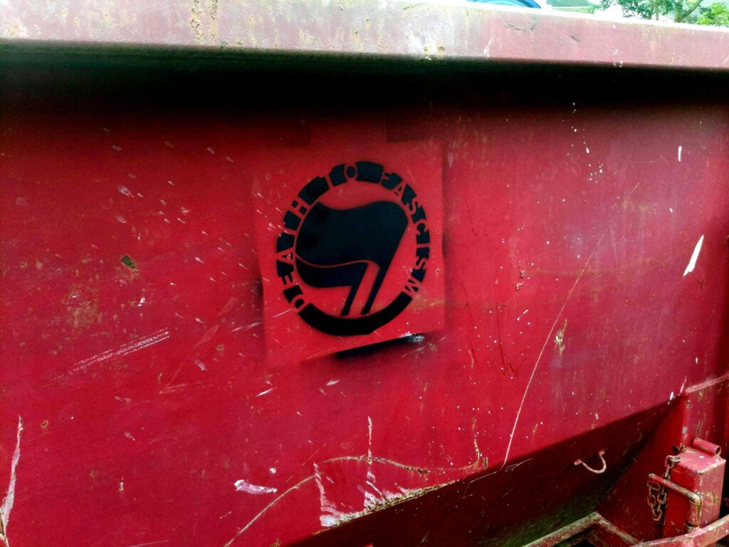 The side of a red dumpster with a black stenciled painting of antifascist flags with the words "Death to Fascism" arced above them.