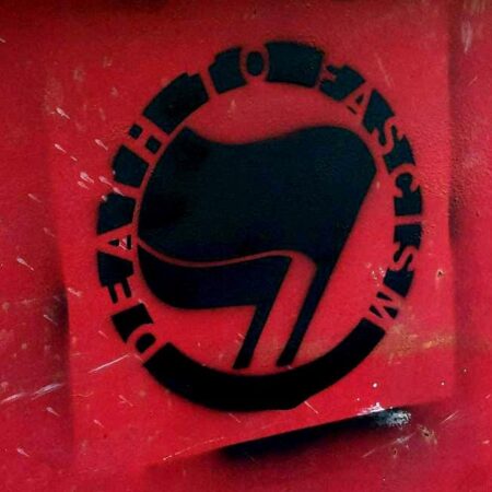 The side of a red dumpster with a black stenciled painting of antifascist flags with the words "Death to Fascism" arced above them.