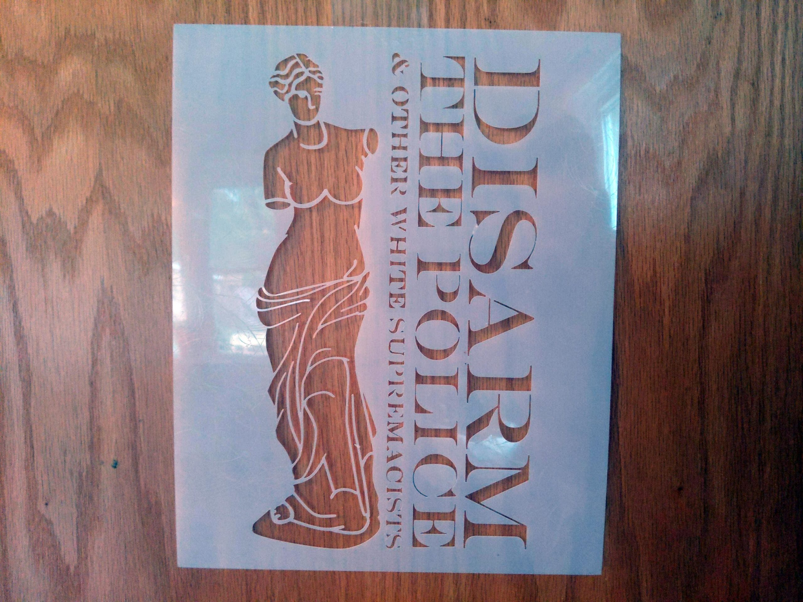 A woodgrain background upon which a stencil is laid. The translucent mylar stencil depicts a line drawing of the Venus de Milo sculpture which is characteristically armless. She abuts the words "Disarm the Police & other White Supremacists."