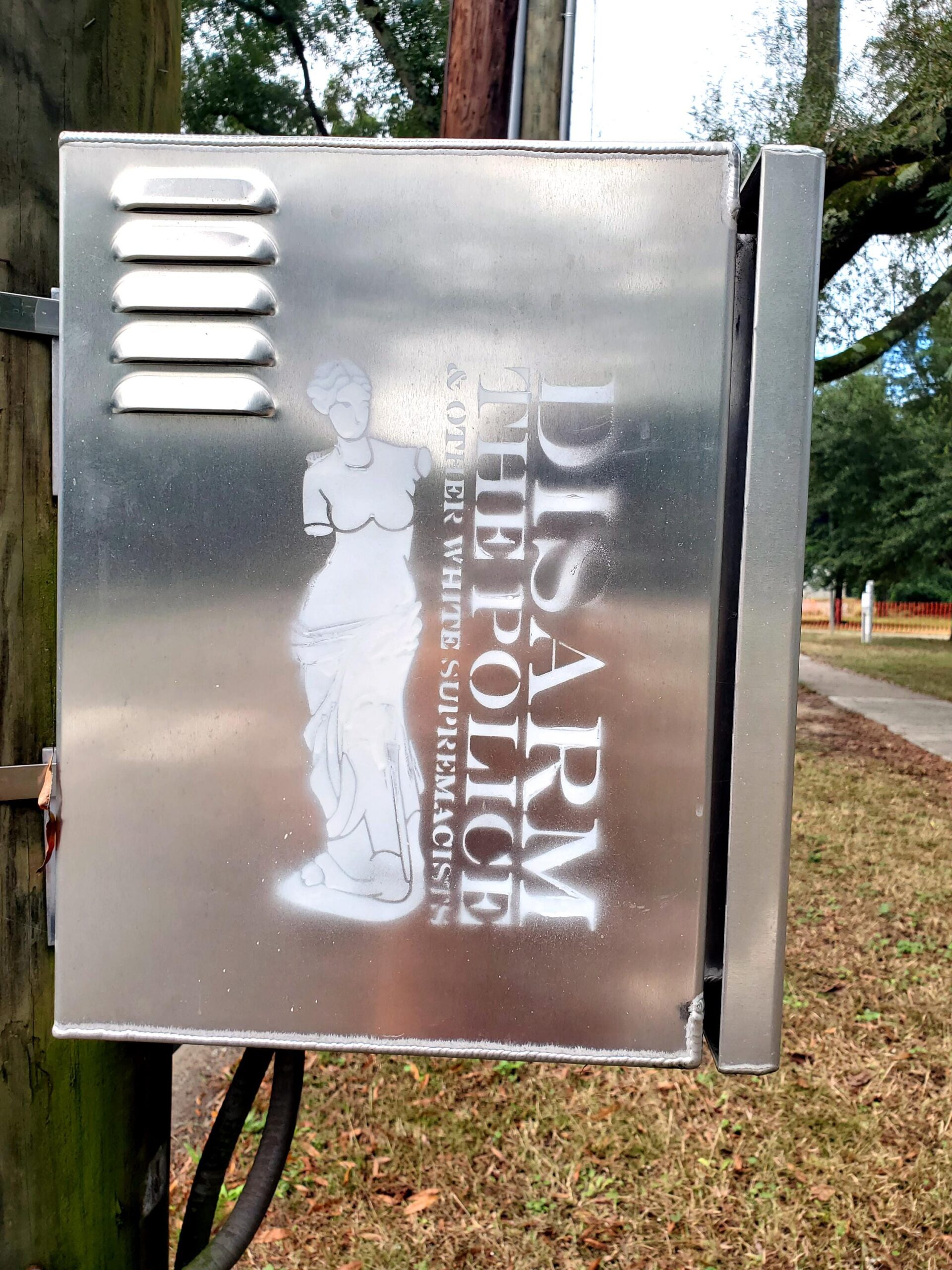 A metallic electrical box is painted with a stencil line drawing of the Venus de Milo sculpture which is characteristically armless. She abuts the words "Disarm the Police & other White Supremacists."
