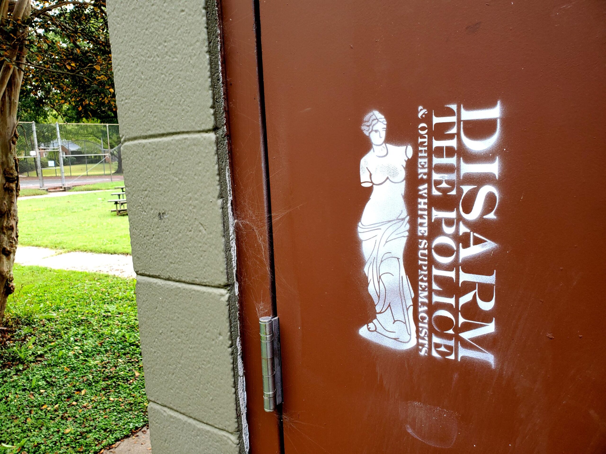 A brown door on a cinderblock structure in a grassy area. The door is painted with a line drawing of the Venus de Milo sculpture which is characteristically armless. She abuts the words "Disarm the Police & other White Supremacists."