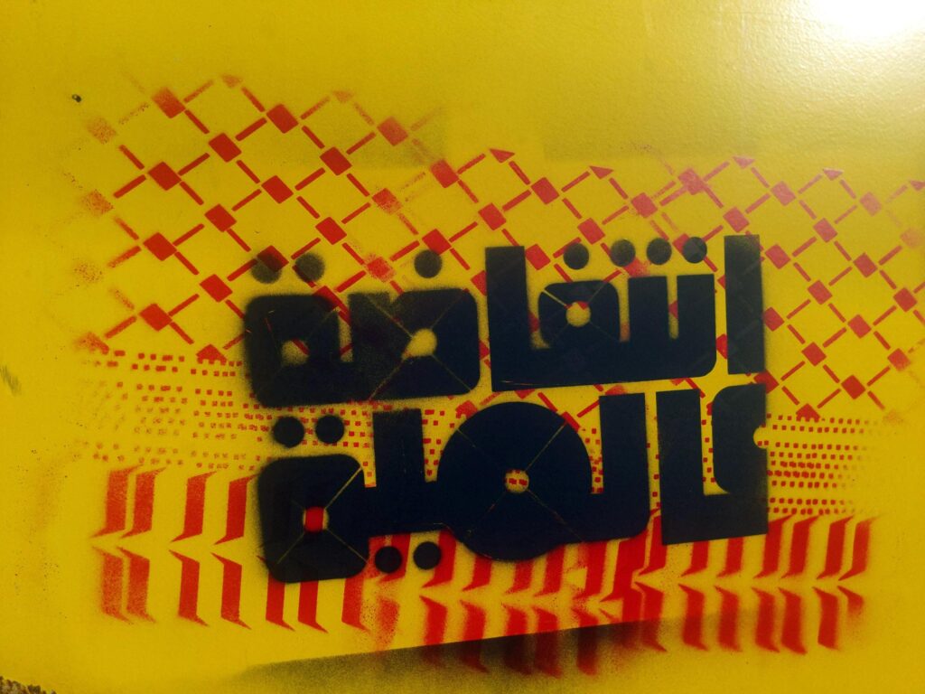 A yellow surface on which someone has spraypainted a 2-layer stencil piece, with the background of keffiyeh embroidery and a foreground of "نتفاضة عالمية" or "Global Intifada" in Arabic in a rounded typeface reminiscent of kufic calligraphy or 70s typography.