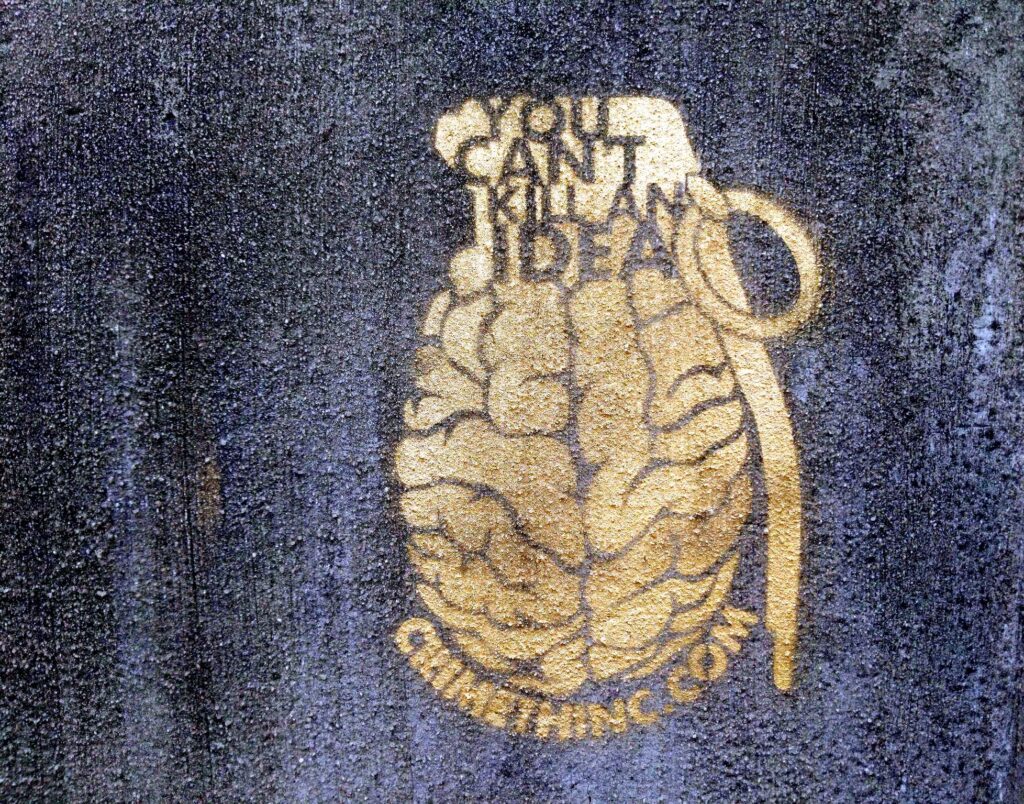 A concrete wall with a stencil spraypainted on it in metallic gold: The stencil reads: "You Can't Kill An Idea" on the fuze of a grenade with a brain for a charge and body. The url crimethinc.com arcs underneath the brainade.
