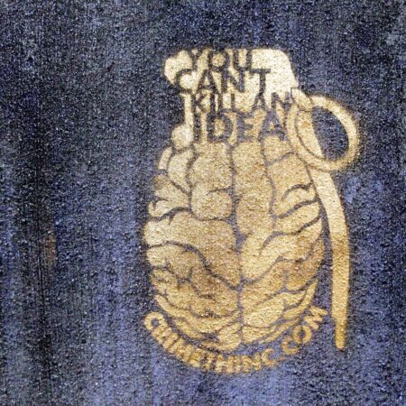A concrete wall with a stencil spraypainted on it in metallic gold: The stencil reads: "You Can't Kill An Idea" on the fuze of a grenade with a brain for a charge and body. The url crimethinc.com arcs underneath the brainade.