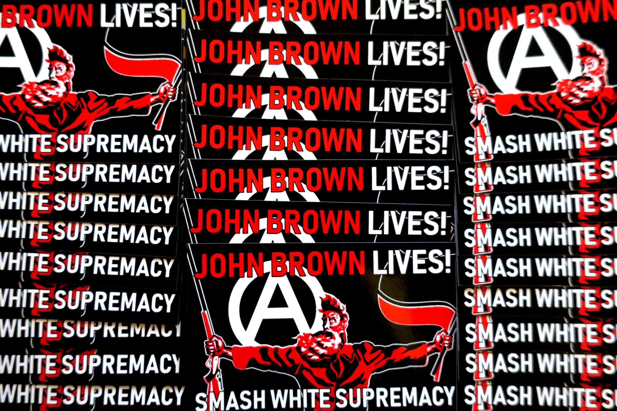 Three tidy columns numbering a total of 25 black stickers in which a three color portrait of John Brown taken from the painting "Tragic Prelude" stands in the center. He holds a Sharps rifle in his right hand and antifascist red and black flags in his left hand. Over his right shoulder looms a large circled A. At the top of the sticker, the words John Brown Lives! in red with the last word white. Across the bottom, the words Smash White Supremacy in white.