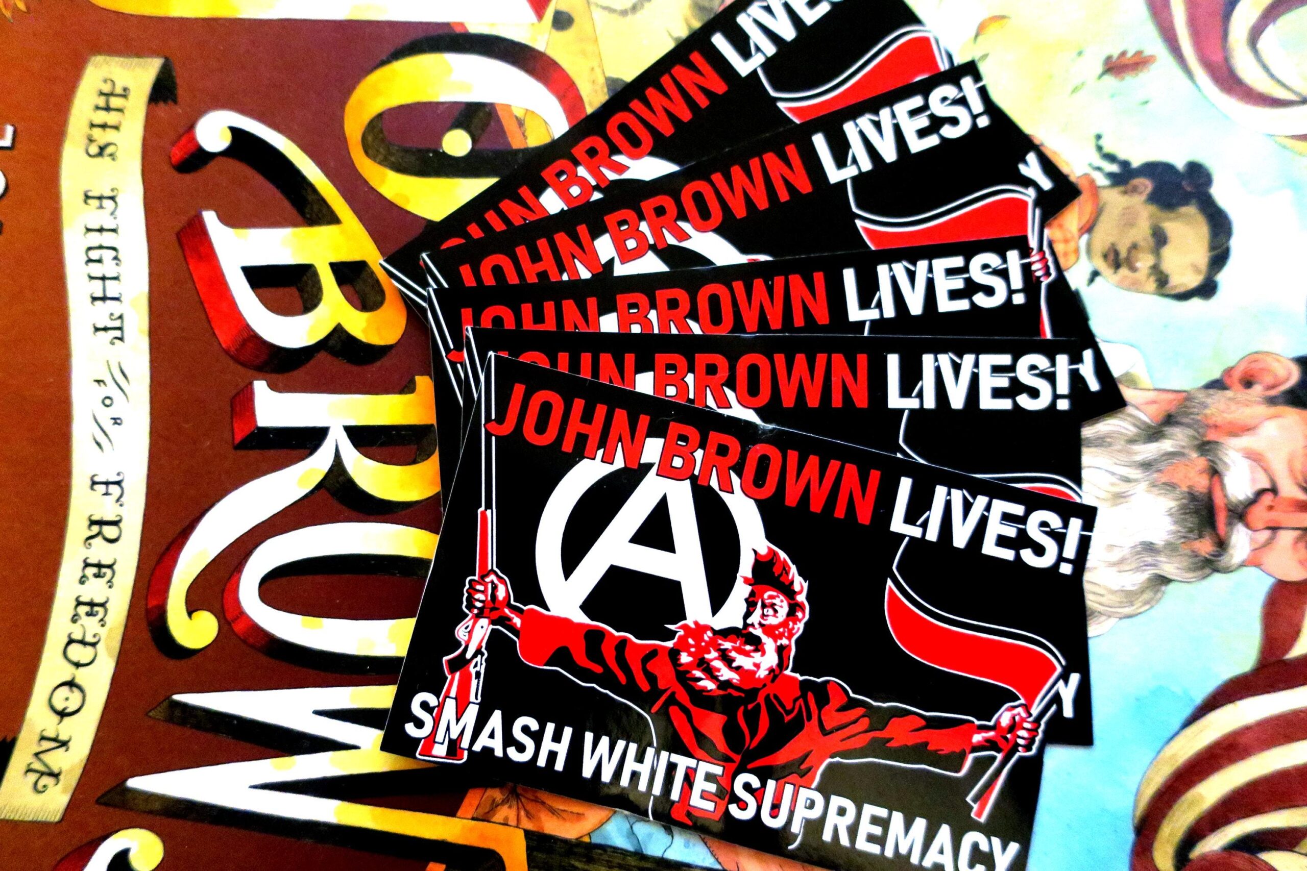 On top of the book jacket for John Brown: His Fight for Freedom, a fan shaped arrangement of 5 black stickers in which a three color portrait of John Brown taken from the painting "Tragic Prelude" stands in the center. He holds a Sharps rifle in his right hand and antifascist red and black flags in his left hand. Over his right shoulder looms a large circled A. At the top of the sticker, the words John Brown Lives! in red with the last word white. Across the bottom, the words Smash White Supremacy in white.