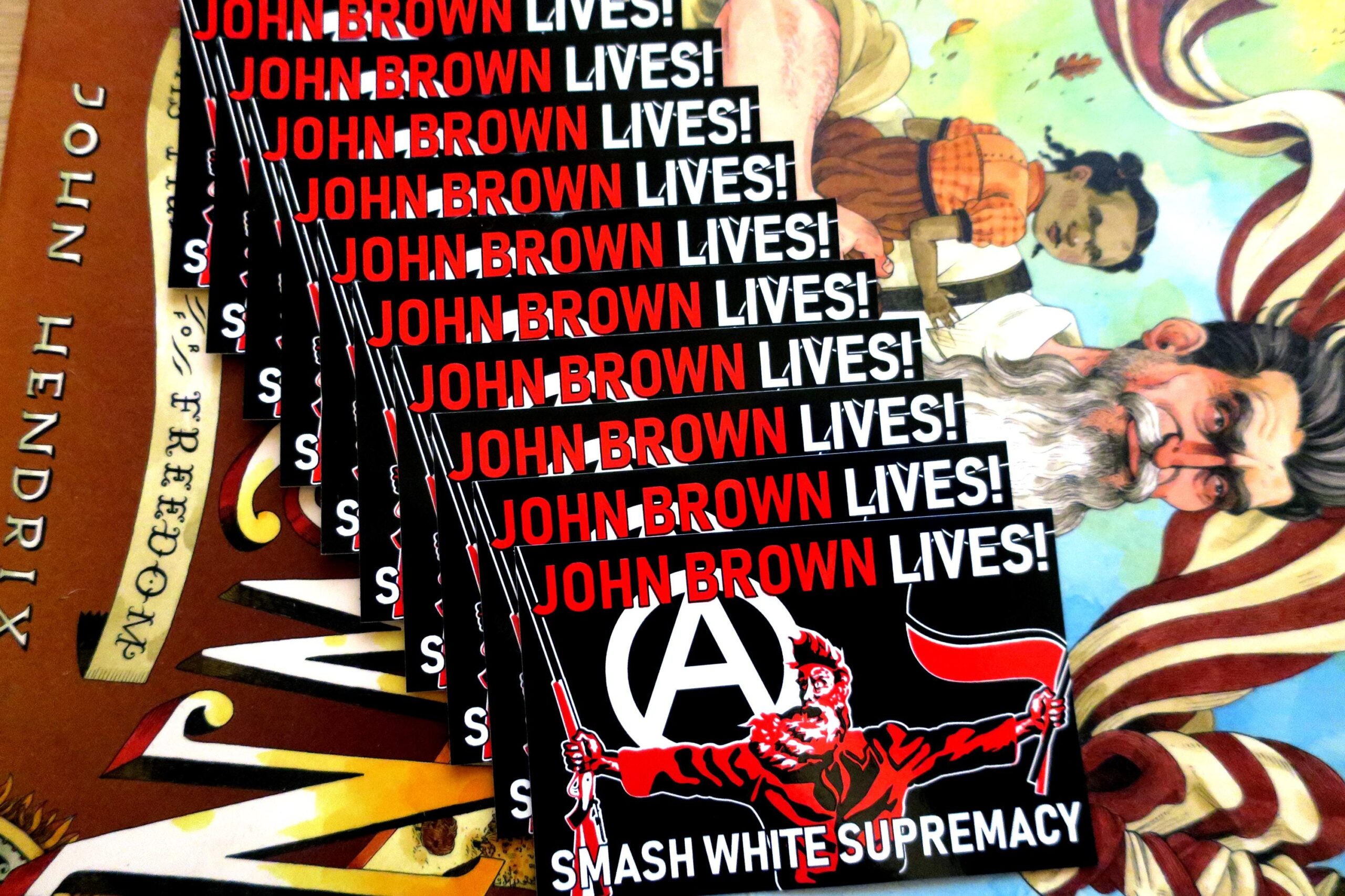 On top of the book jacket for John Brown: His Fight for Freedom, a cascade of 10 black stickers in which a three color portrait of John Brown taken from the painting "Tragic Prelude" stands in the center. He holds a Sharps rifle in his right hand and antifascist red and black flags in his left hand. Over his right shoulder looms a large circled A. At the top of the sticker, the words John Brown Lives! in red with the last word white. Across the bottom, the words Smash White Supremacy in white.