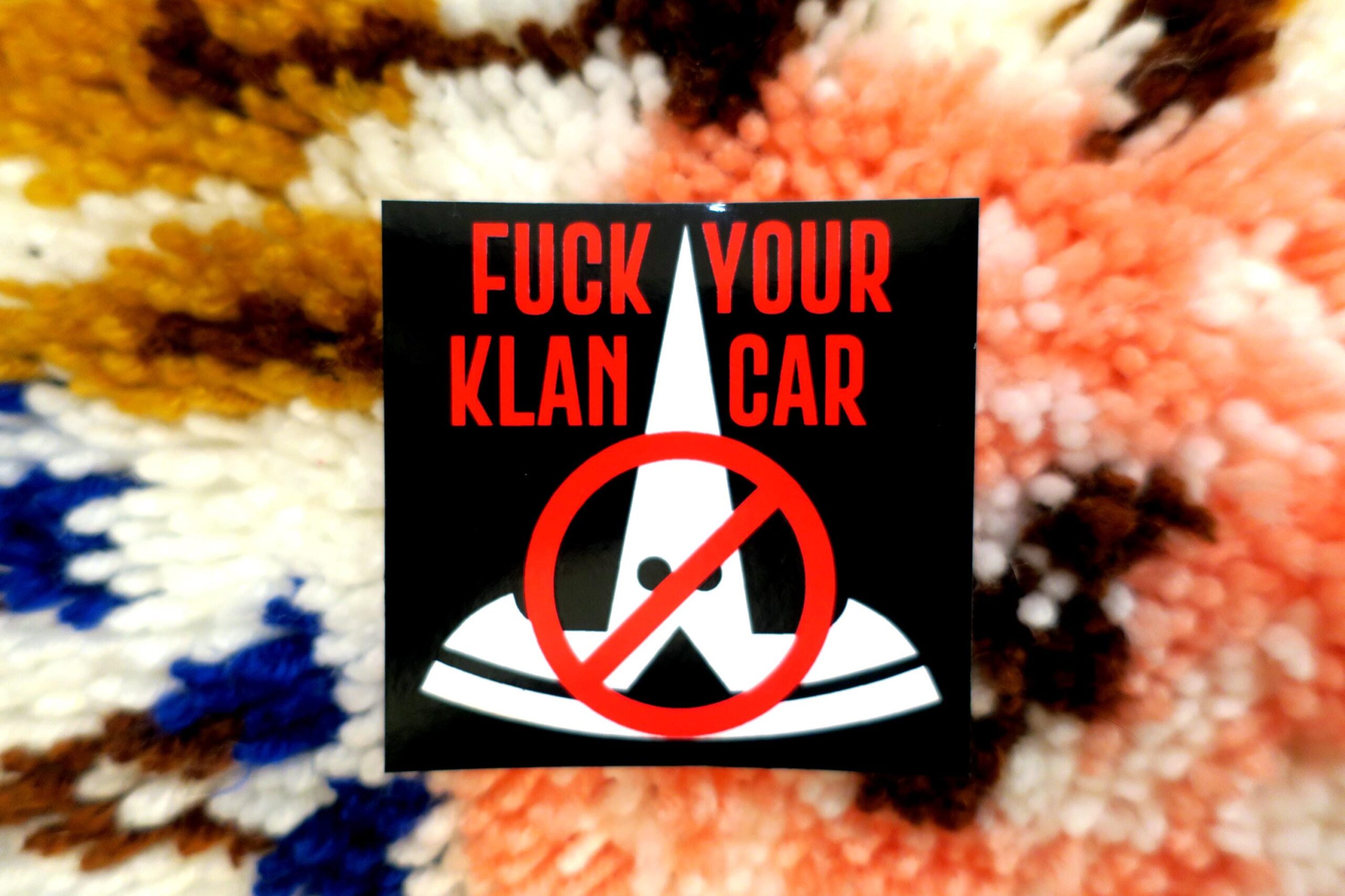 On a colorful high pile carpet, a black sticker: The tesla logo in white, turned upside down and given stupid little eyes looks like a fucking klansman. This is crossed out with the words "FUCK YOUR KLAN CAR" in red across the top.