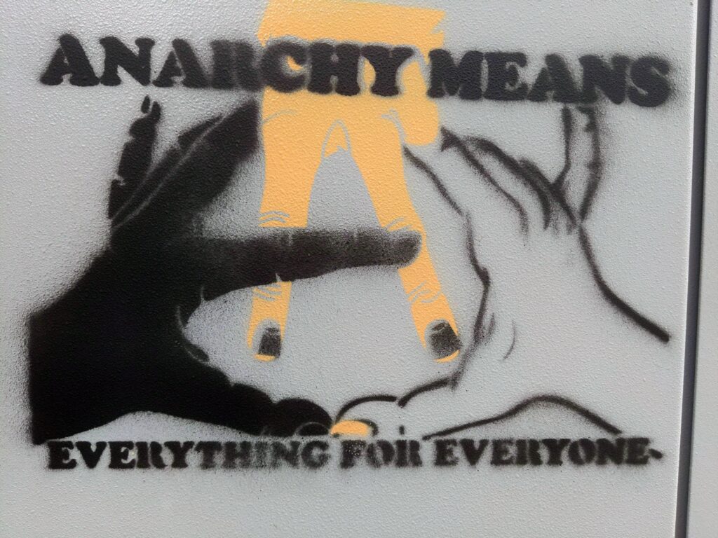 The grey textured surface of an electrical box, spraypainted with black and orangey tan: "Anarchy Means" above three intertwined hands of varying skin tones that compose a circled A. Below, the words, "Everything for Everyone."