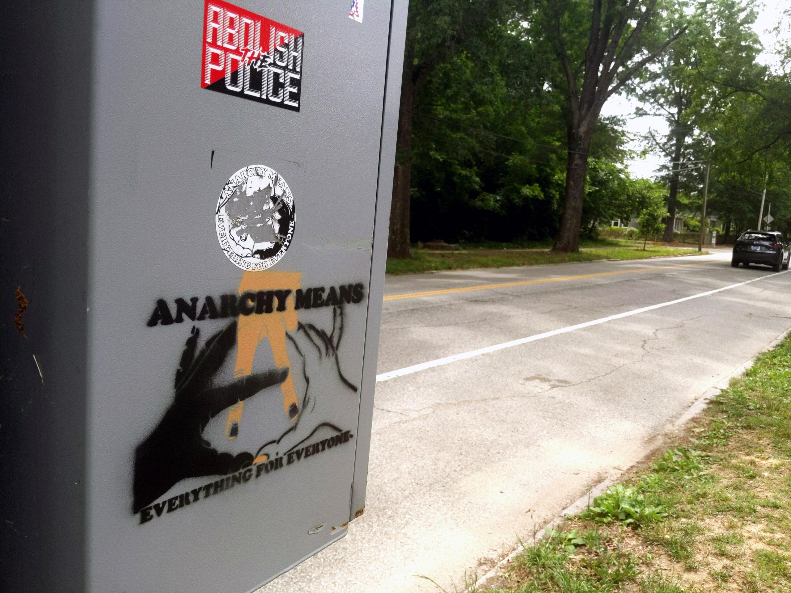 On a tree-lined street, a grey electrical box, with two stickers affixed to it. Below these, it has been spraypainted with black and orangey tan with a stencil: "Anarchy Means" above three intertwined hands of varying skin tones that compose a circled A. Below, the words, "Everything for Everyone."