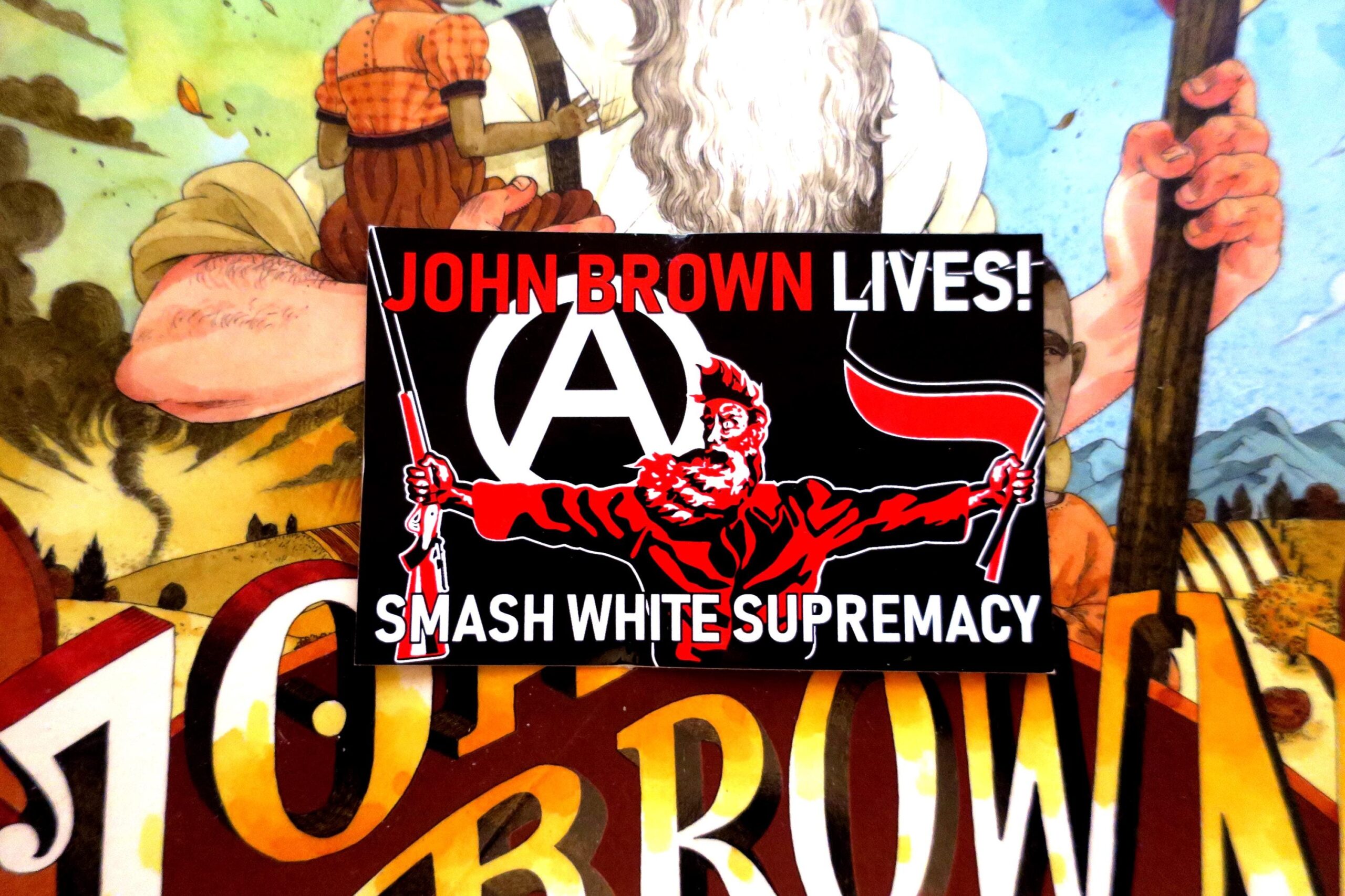 On top of the book jacket for John Brown: His Fight for Freedom, a black sticker in which a three color portrait of John Brown taken from the painting "Tragic Prelude" stands in the center. He holds a Sharps rifle in his right hand and antifascist red and black flags in his left hand. Over his right shoulder looms a large circled A. At the top of the sticker, the words John Brown Lives! in red with the last word white. Across the bottom, the words Smash White Supremacy in white.
