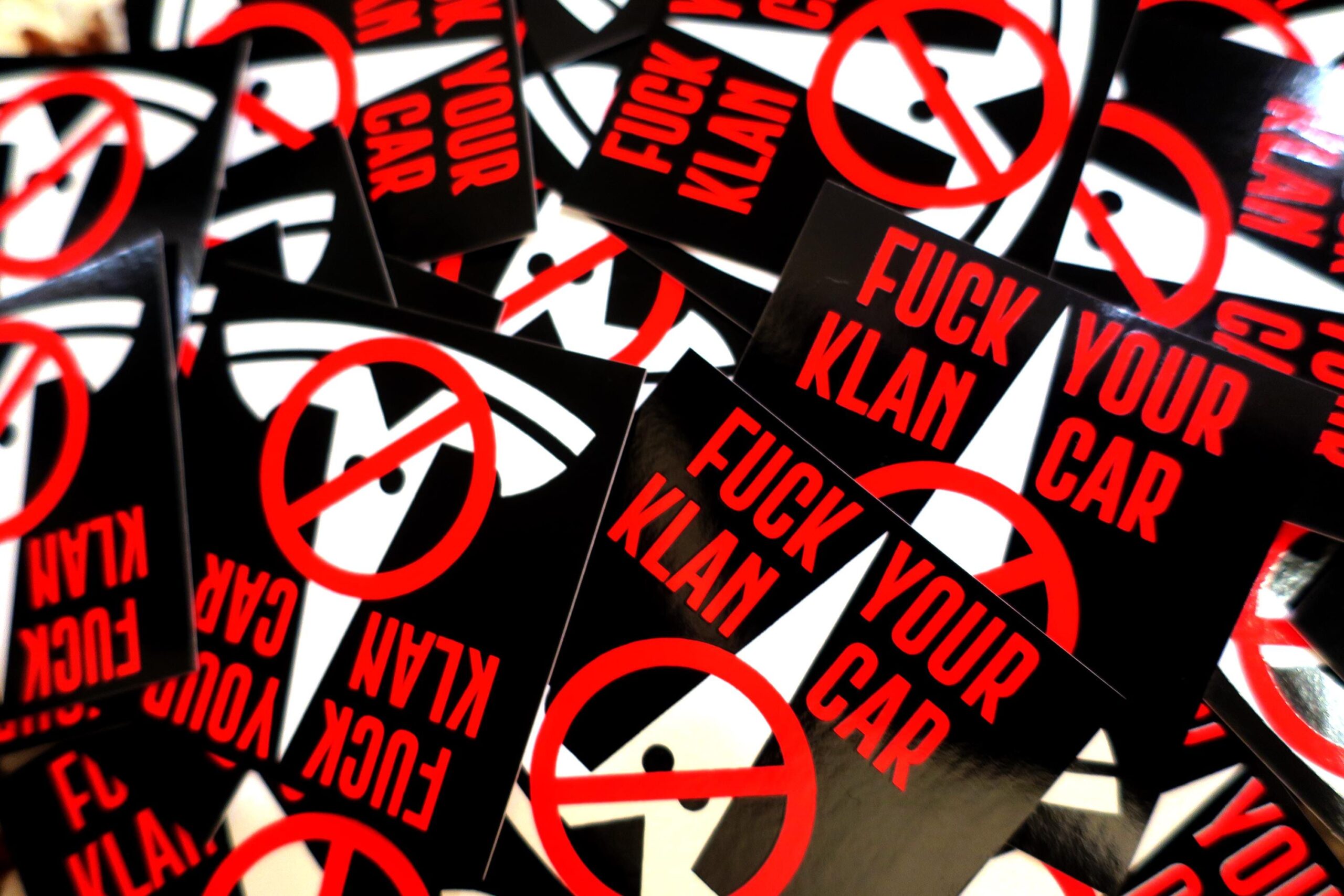 A messy pile of black stickers: The tesla logo in white, turned upside down and given stupid little eyes looks like a fucking klansman. This is crossed out with the words "FUCK YOUR KLAN CAR" in red across the top.