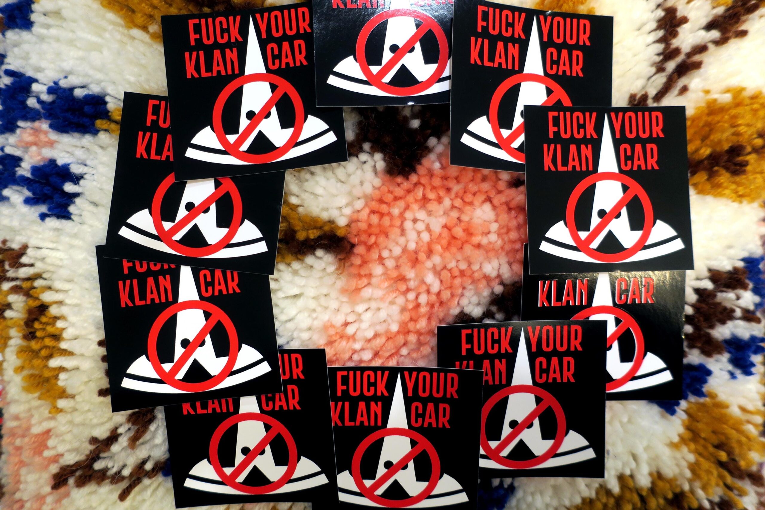 On a colorful high pile carpet, 10 black stickers in a circle: The tesla logo in white, turned upside down and given stupid little eyes looks like a fucking klansman. This is crossed out with the words "FUCK YOUR KLAN CAR" in red across the top.