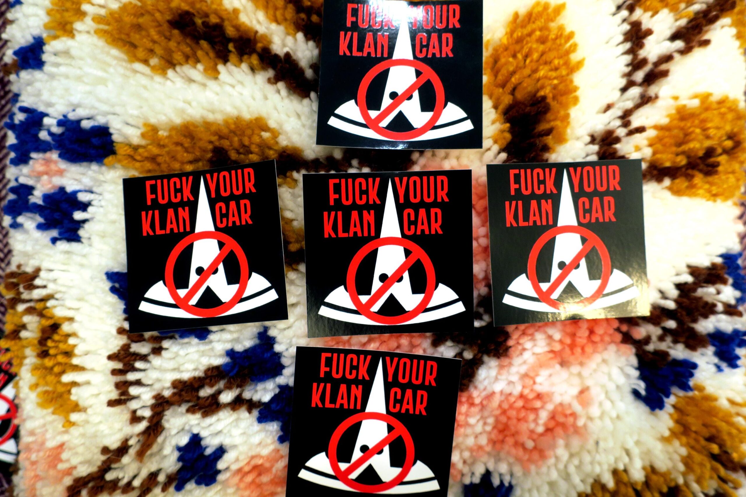 On a colorful high pile carpet, a cross of 5 black stickers: The tesla logo in white, turned upside down and given stupid little eyes looks like a fucking klansman. This is crossed out with the words "FUCK YOUR KLAN CAR" in red across the top.