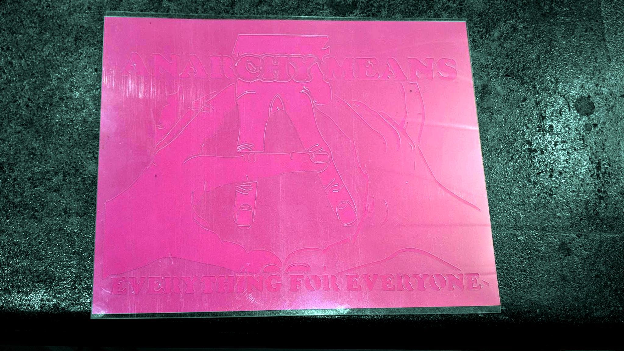 A transparent mylar stencil atop a pink piece of paper, itself resting on smooth concrete. The stencil reads: "Anarchy Means" above three intertwined hands that compose a circled A. Below, the words, "Everything for Everyone."