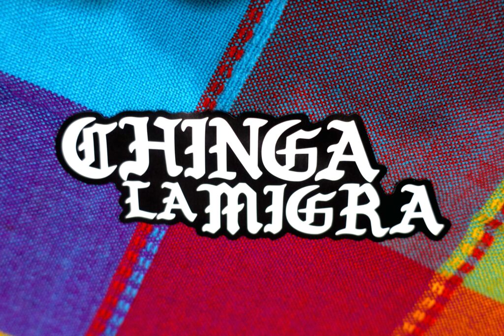 Against a brightly colored knit facbric, a black and white sticker reading Chinga La Migra in simple blackletter typeface white, with a thick black outline (not a border).