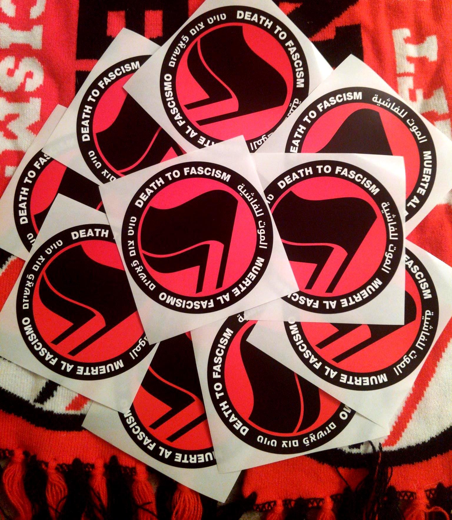 On a backdrop of knit antifa scarves, 10 stickers in a messy pile show a thick black circle containing the words: "Death to Fascism, Muerte al Fascismo, الموت للفاشية, טויט צום פֿאַשיזם, ." Inside this circle are two black flags arranged over a red field recognizable as the antifascist logo.
