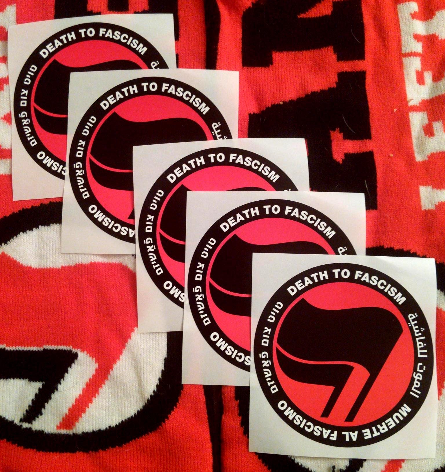 On a backdrop of knit antifa flags, 5 stickers in a cascading pattern show a thick black circle containing the words: "Death to Fascism, Muerte al Fascismo, الموت للفاشية, טויט צום פֿאַשיזם, ." Inside this circle are two black flags arranged over a red field recognizable as the antifascist logo.