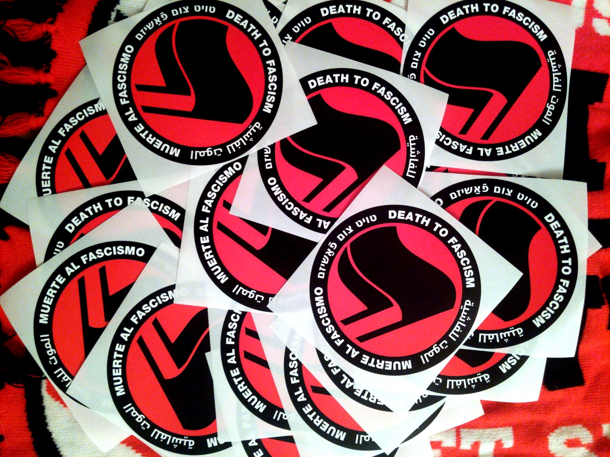 On a backdrop of knit antifa scarves, 25 stickers in a messy pile show a thick black circle containing the words: "Death to Fascism, Muerte al Fascismo, الموت للفاشية, טויט צום פֿאַשיזם, ." Inside this circle are two black flags arranged over a red field recognizable as the antifascist logo.