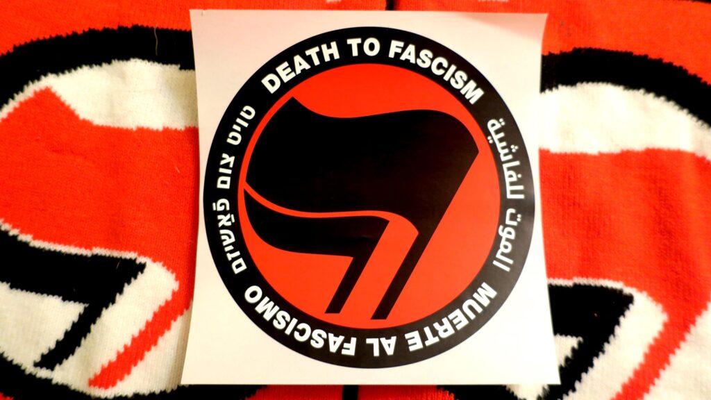 On a backdrop of knit antifa flags, a red, black and white sticker with a thick black circle containing the words: "Death to Fascism, Muerte al Fascismo, الموت للفاشية, טויט צום פֿאַשיזם, ." Inside this circle are two black flags arranged over a red field recognizable as the antifascist logo.