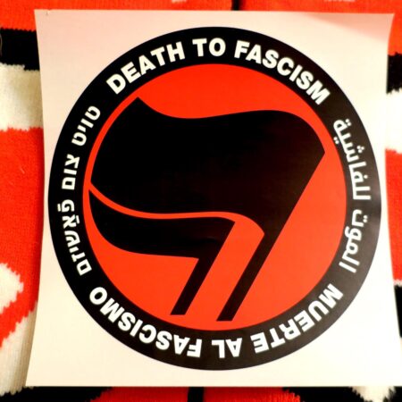 On a backdrop of knit antifa flags, a red, black and white sticker with a thick black circle containing the words: "Death to Fascism, Muerte al Fascismo, الموت للفاشية, טויט צום פֿאַשיזם, ." Inside this circle are two black flags arranged over a red field recognizable as the antifascist logo.