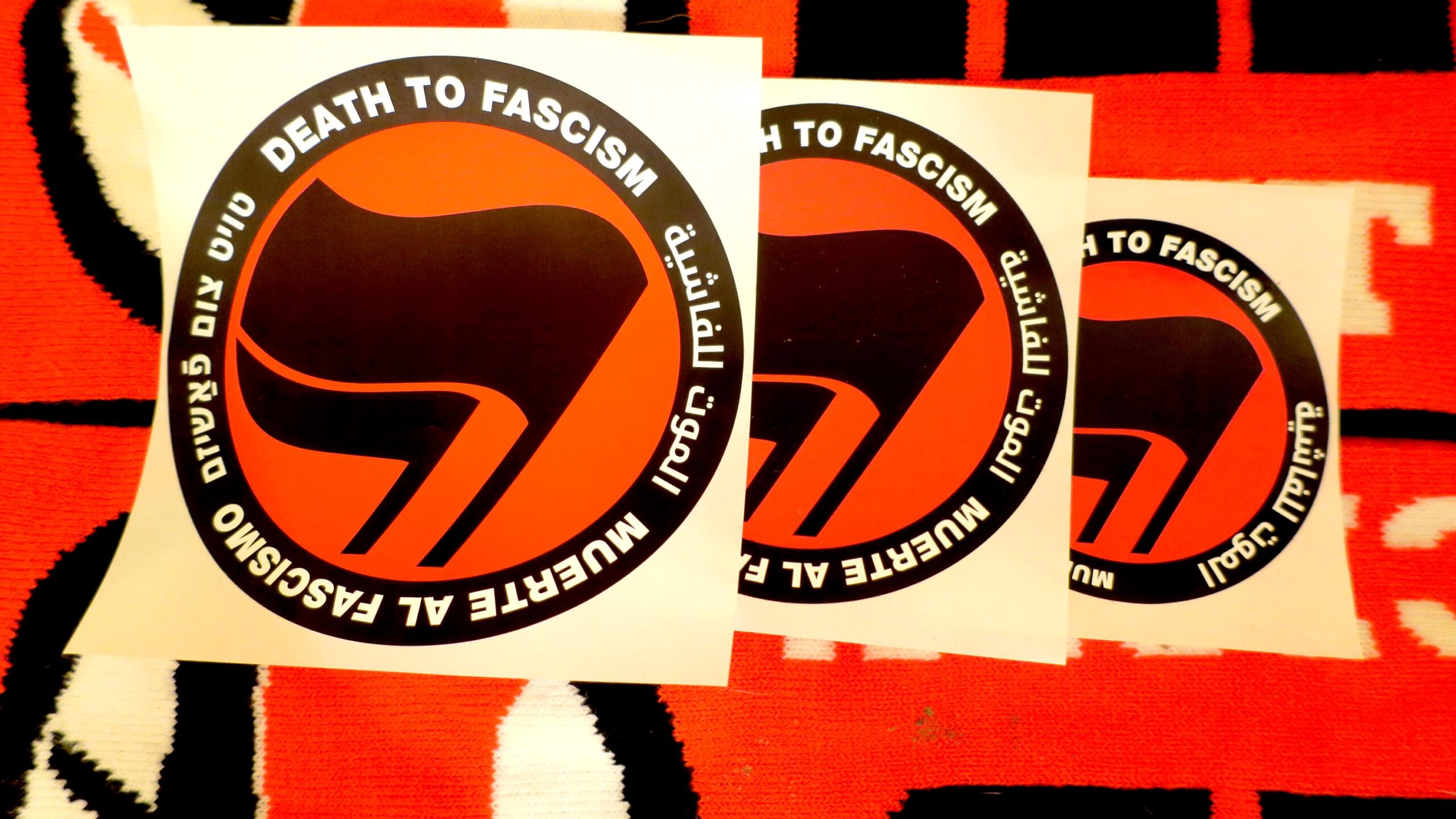 On a backdrop of knit antifa flags, 3 stickers in descending size representing the sticker's design revisions show a thick black circle containing the words: "Death to Fascism, Muerte al Fascismo, الموت للفاشية, טויט צום פֿאַשיזם, ." Inside this circle are two black flags arranged over a red field recognizable as the antifascist logo.