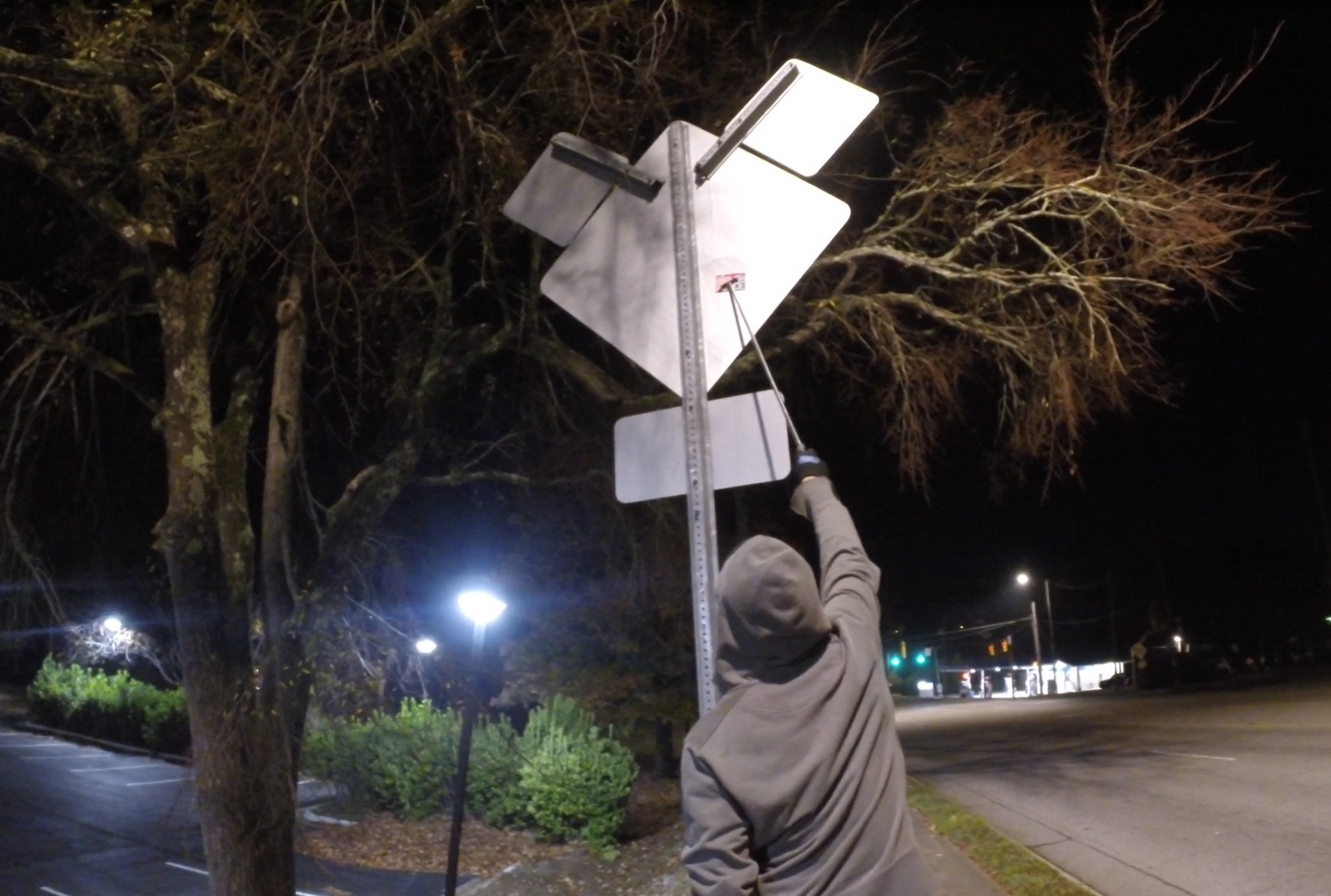 A hooded figure reaches up towards a traffic sign with a stick em up pole extending their reach to put a sticker on the back, high up!