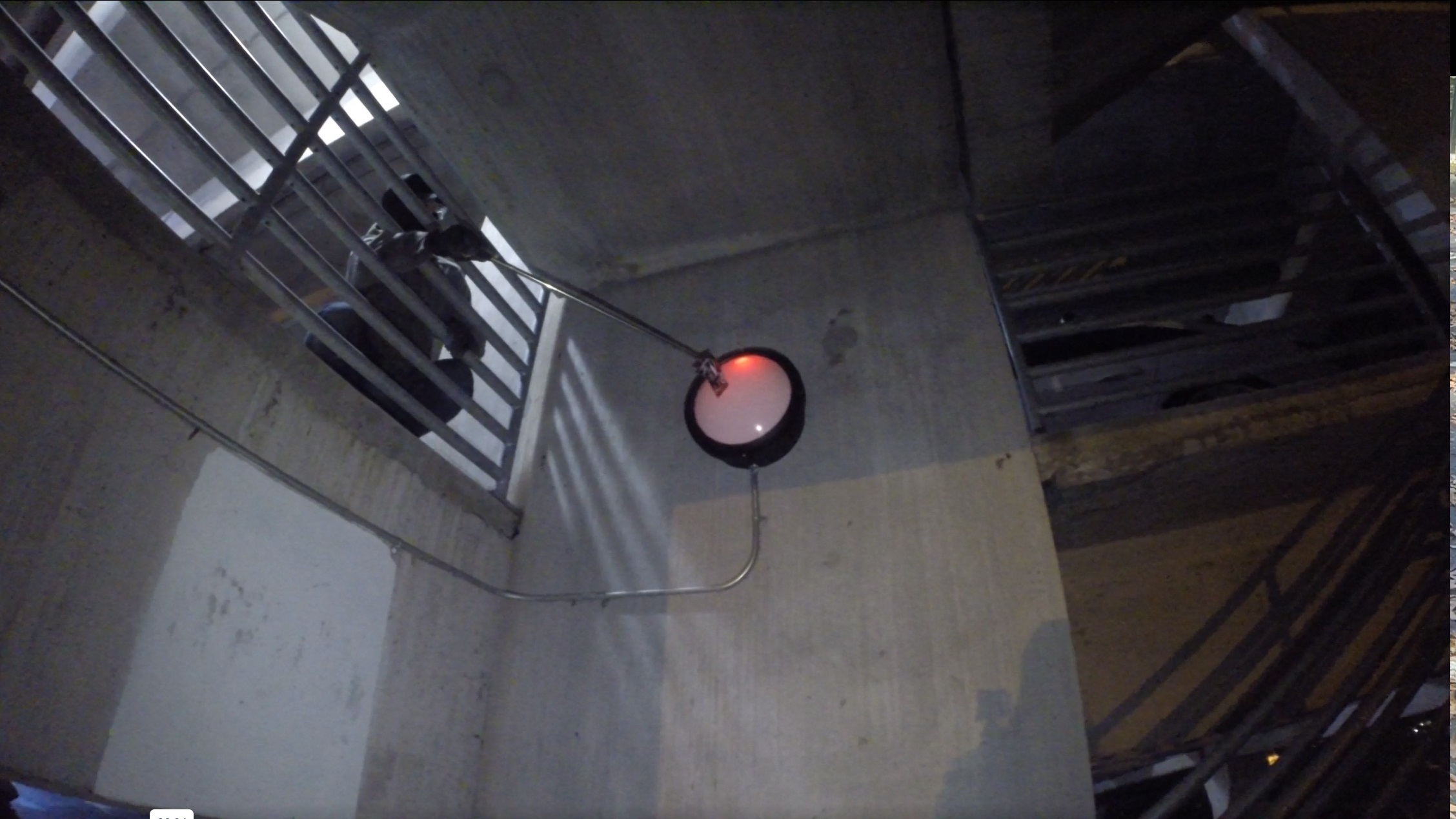 A hooded figure reaches through a barred grate with a stick em up pole extending their reach to put a sticker in the center of a tall (and far!) light fixture