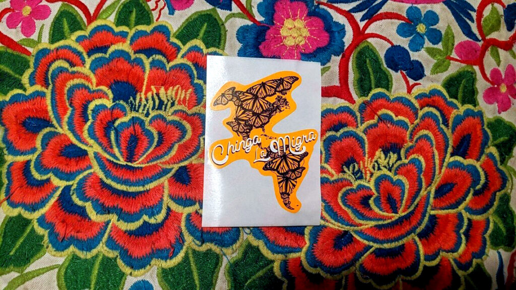 Atop a colorfully embroidered pillow, a sticker of an orange shape of North America and South America filled with monarch butterflies. The words Chinga La Migra swoop across the continents in white, black-shadowed script.