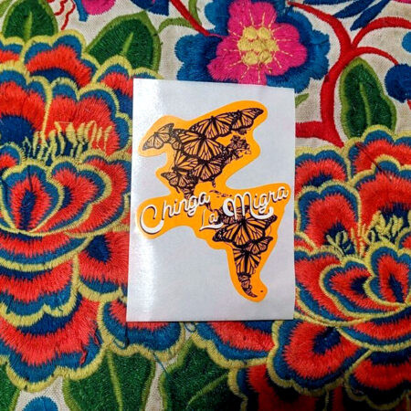 Atop a colorfully embroidered pillow, a sticker of an orange shape of North America and South America filled with monarch butterflies. The words Chinga La Migra swoop across the continents in white, black-shadowed script.