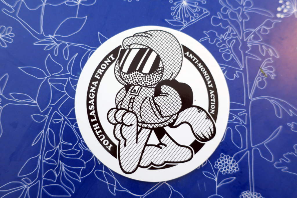 Background of a blue field with white outlines of flowers and leaves. In the foreground, a white circular sticker. Inside the white circles is a black circle with the Words "Youth Lasagna Front" and "Anti-Monday Action" in Cooper Black typeface, reminiscent of a certain orange cat comic title. In the center, between these two phrases, a hooded, masked figure with sunglasses and a black backpack walks confidently through the frame. The large paws and the striped tail sticking out the back reveal that this is Lasagna Cat.